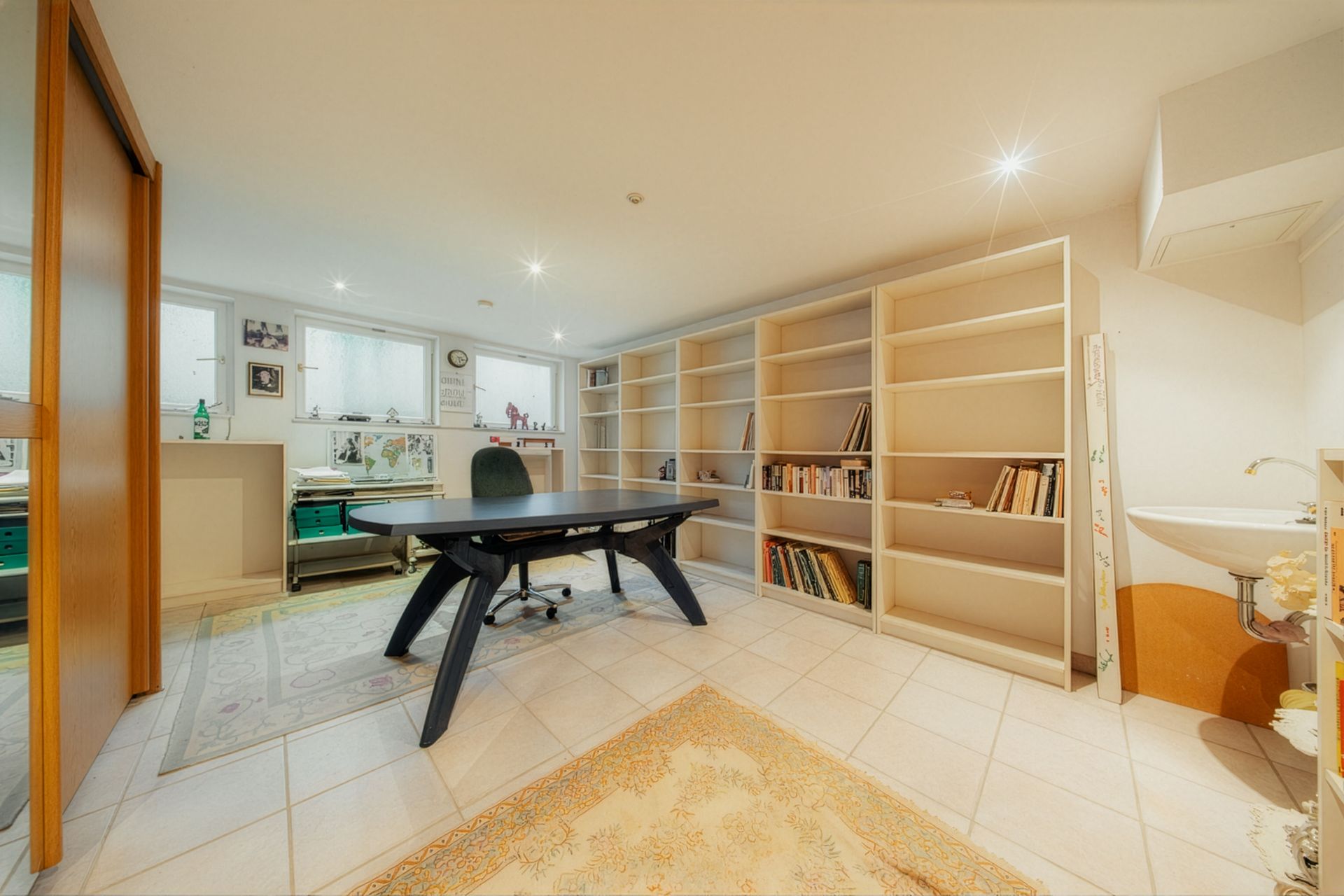 Helles Arbeitszimmer mit großem Schreibtisch, Bücherregalen, Teppich und Waschbecken.