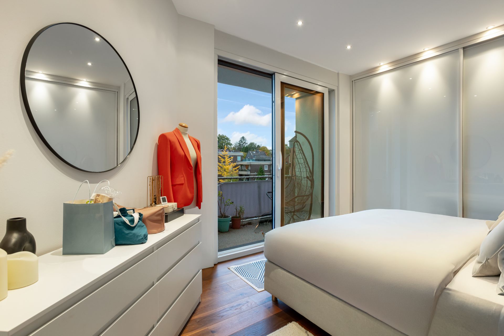 Modernes Schlafzimmer mit Bett, Kommode, rundem Spiegel und Zugang zu einem Balkon.