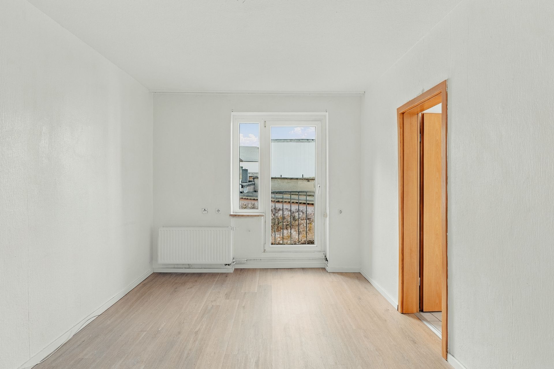 Heller leerer Raum mit Holzboden, weißer Wand, Fenster und Tür zu einem anderen Raum.
