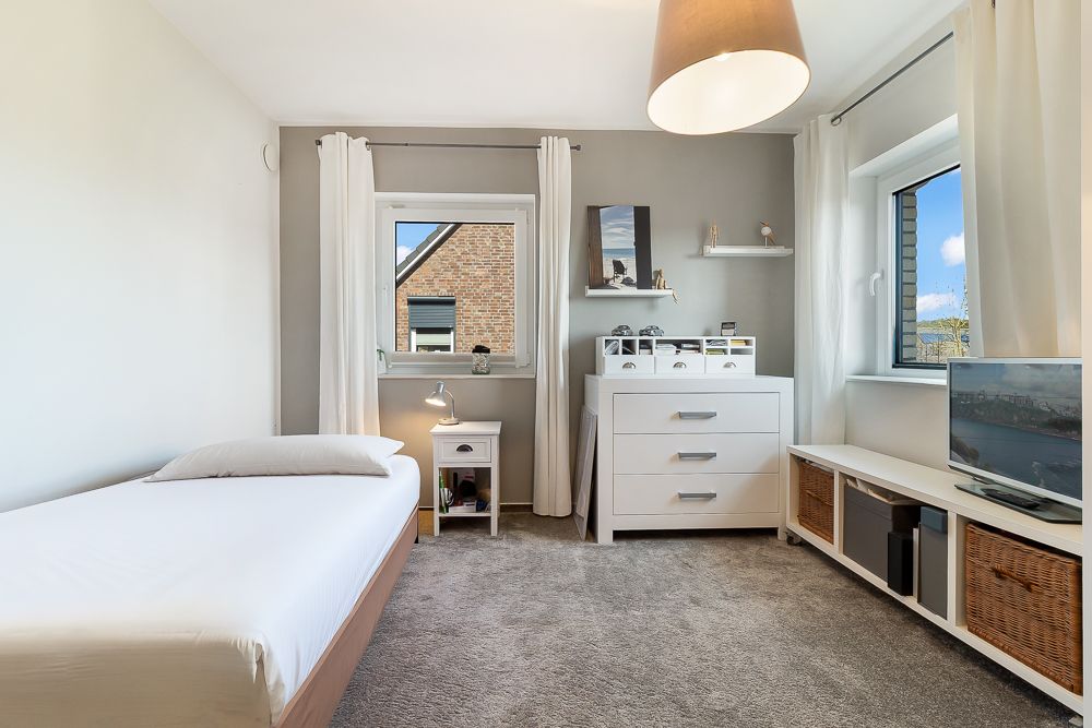 Helles Schlafzimmer mit Einzelbett, Kommode, Fernseher und zwei Fenstern mit weißen Vorhängen.