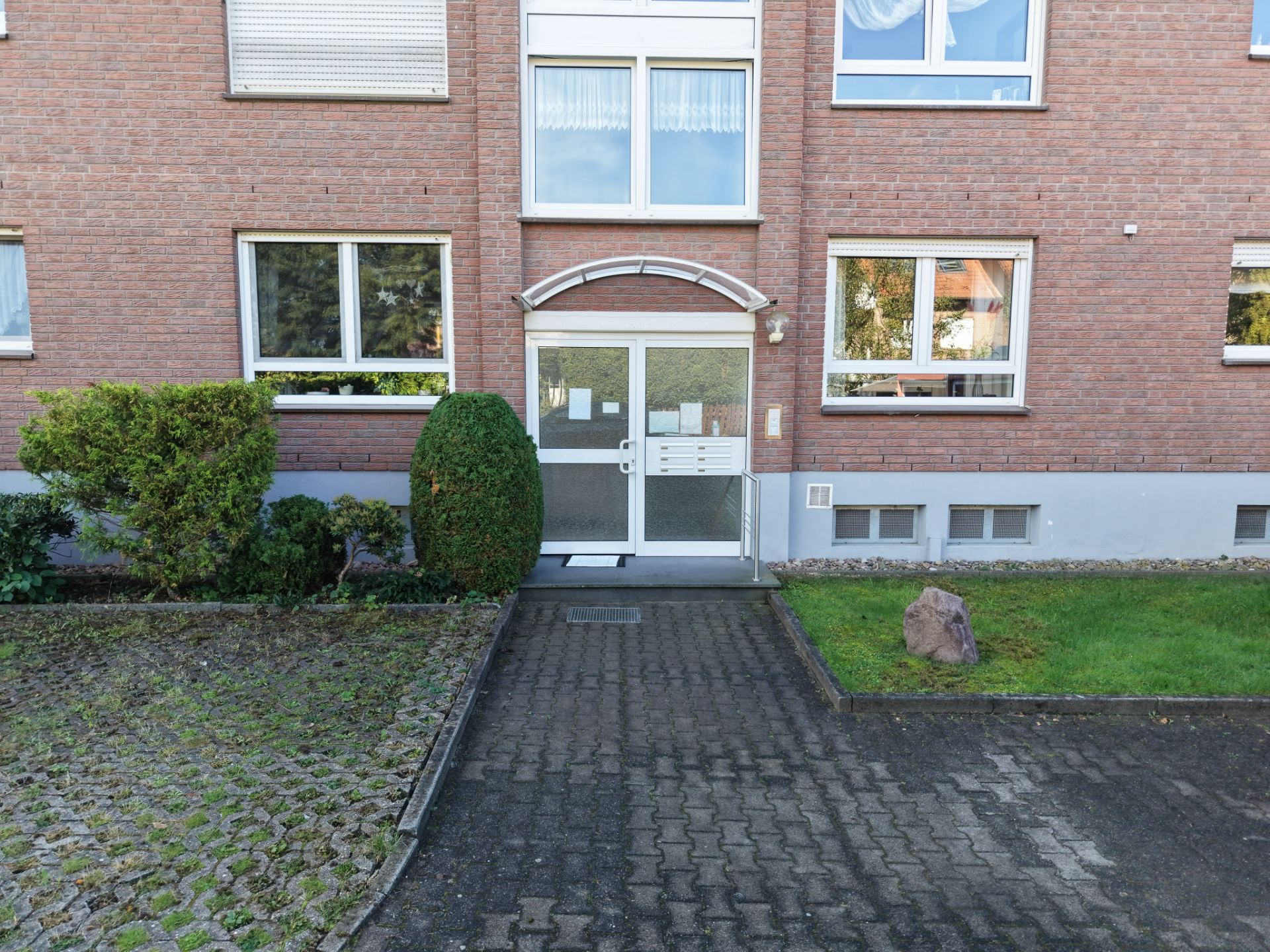 Eingang eines Mehrfamilienhauses mit Glasür und umliegender Bepflanzung.