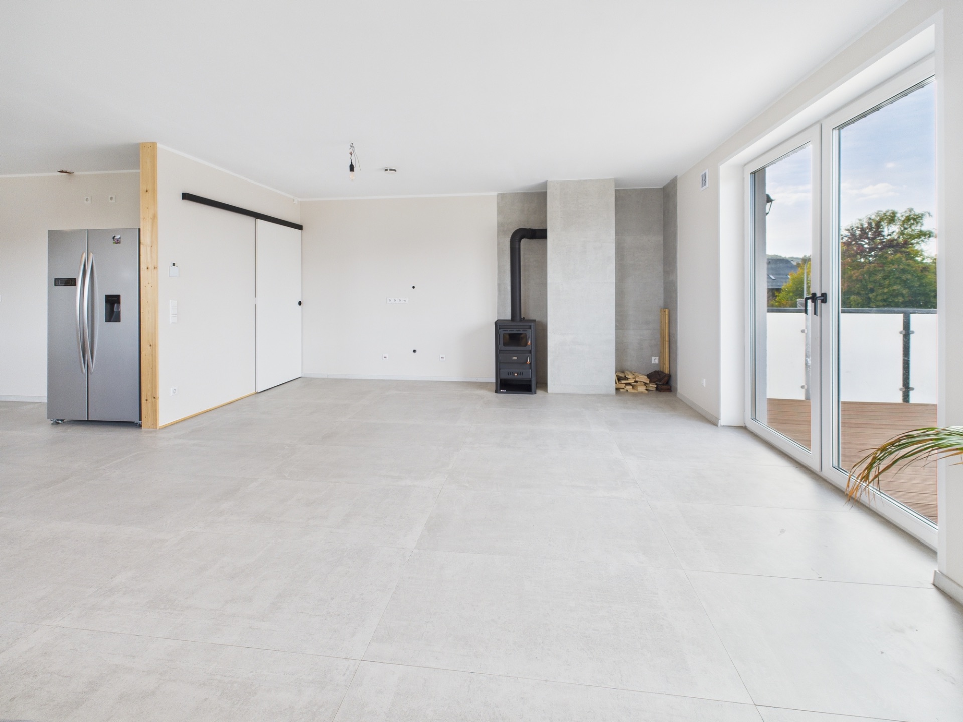 Heller, leerer Raum mit Fliesenboden, Kaminofen, Kühlschrank und großen Fenstern zur Terrasse.