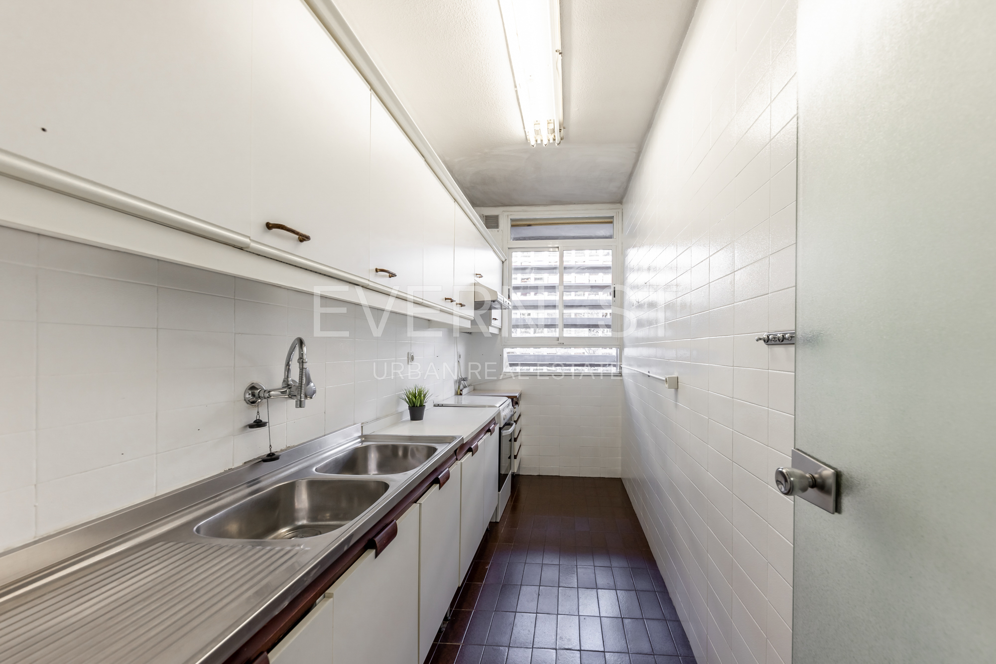 Schmale Küche mit weißen Schränken, Edelstahlspüle und Fenster am Ende des Raums.