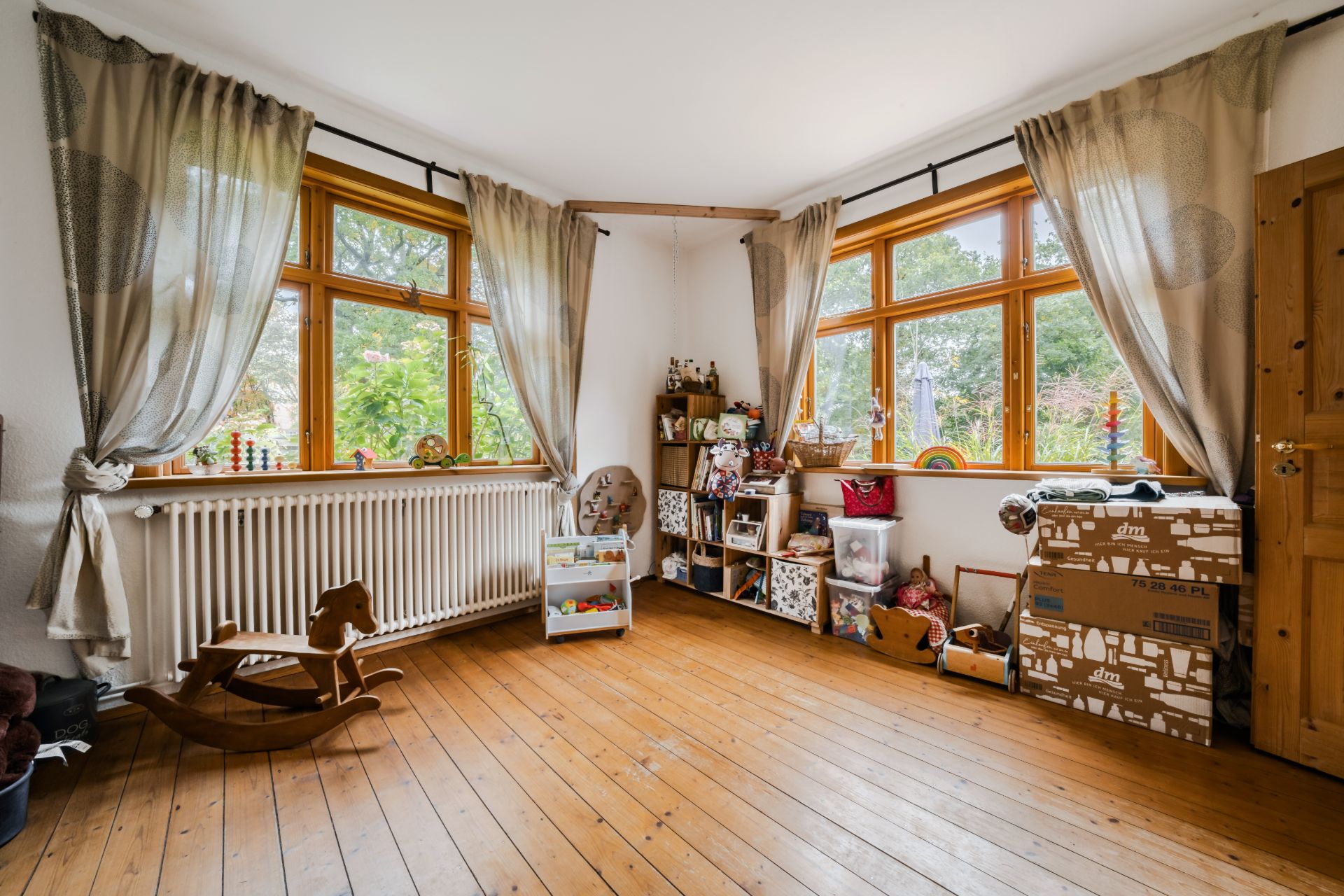 Helles Kinderzimmer mit Holzfußboden, Spielzeugregalen, Vorhängen und großen Fenstern.