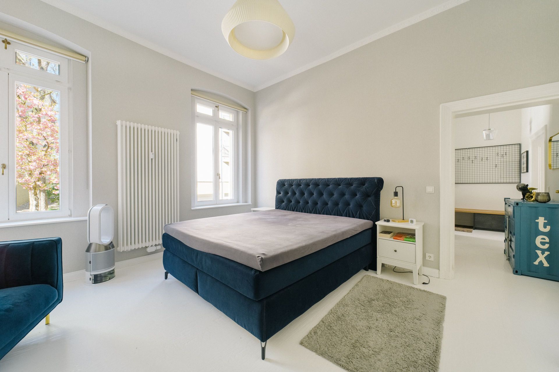 Helles Schlafzimmer mit dunkelblauem Bett, zwei Fenstern, Nachttisch und grauem Teppich.