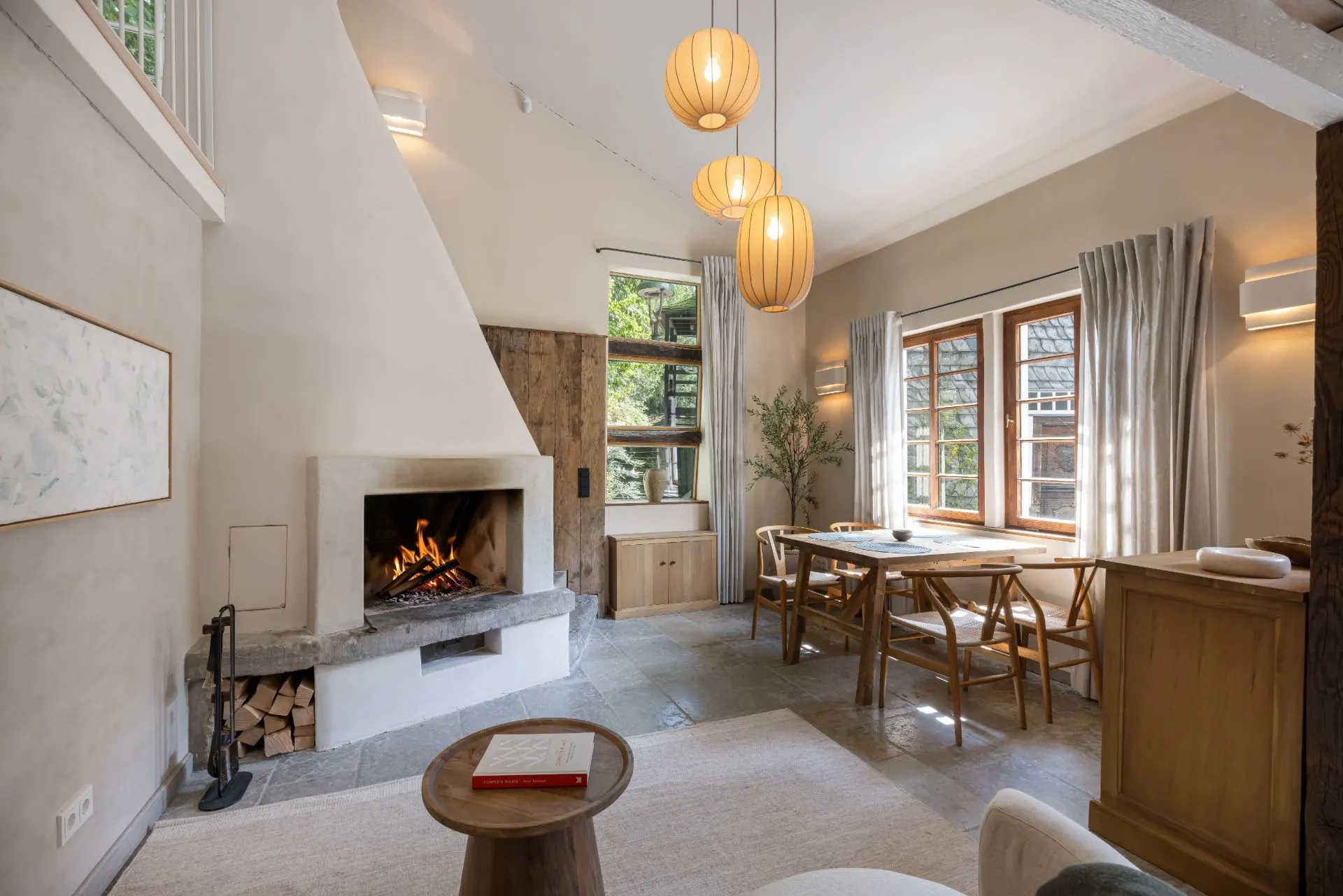 Helles Wohnzimmer mit Kamin, Esstisch, Holzmöbeln und großen Fenstern mit Vorhängen.