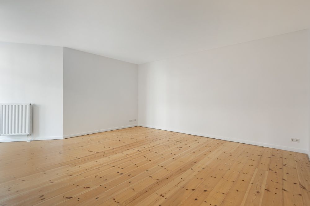 Leerer Raum mit hellem Holzboden, weißen Wänden und einem Heizkörper an der Wand.