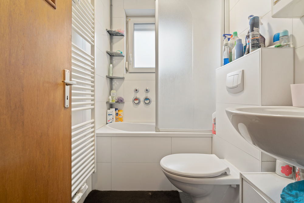 Kleines Badezimmer mit Badewanne, Toilette, Waschbecken und Regalen mit Pflegeprodukten.