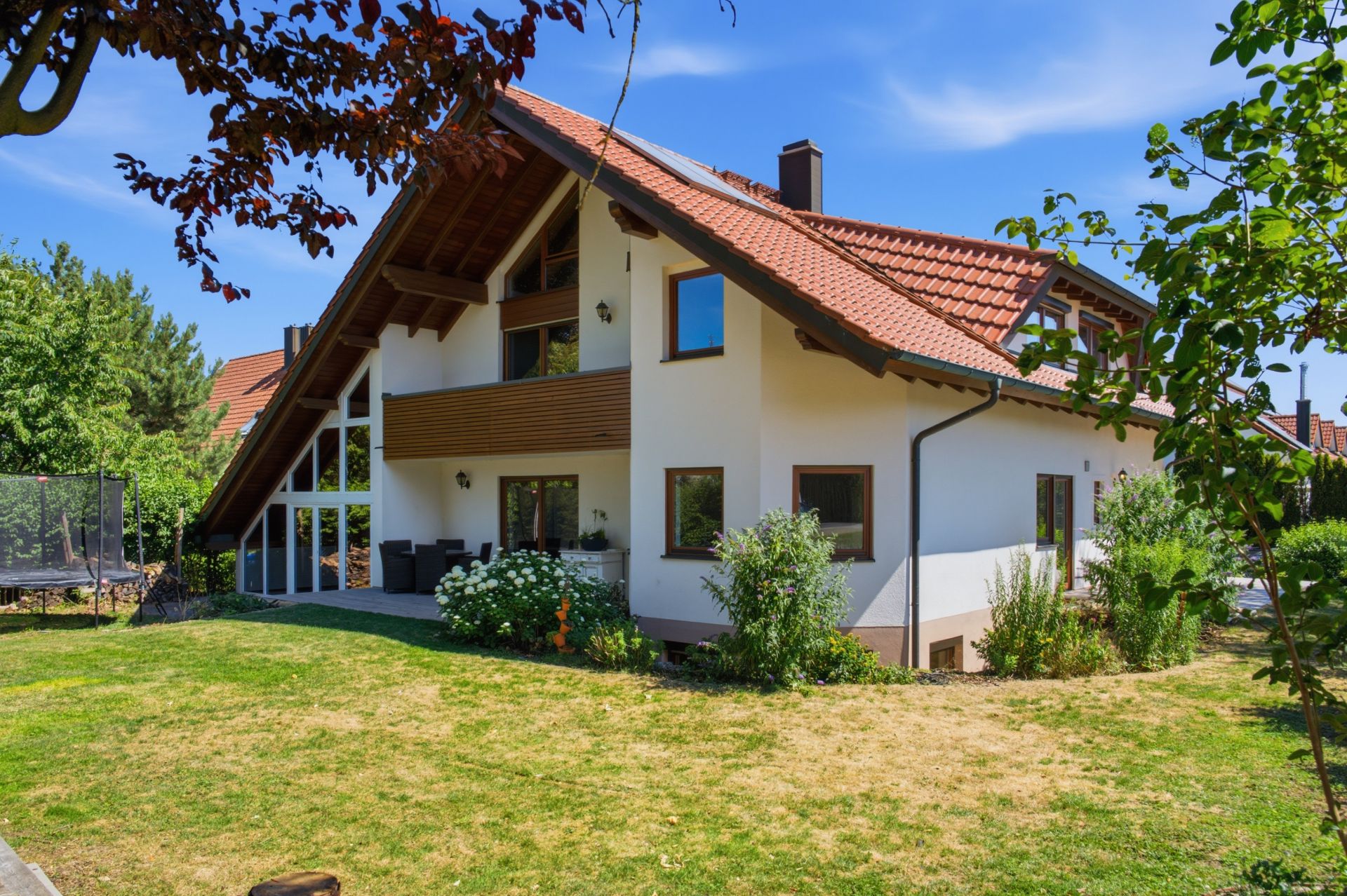 Modernes Einfamilienhaus mit rotem Dach, großem Garten und Terrasse an einem sonnigen Tag.