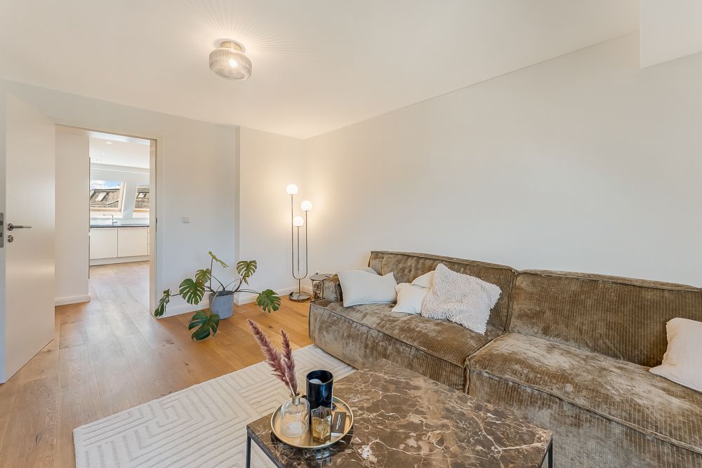 Helles Wohnzimmer mit braunem Sofa, Stehlampe, Pflanze und Zugang zur Küche.
