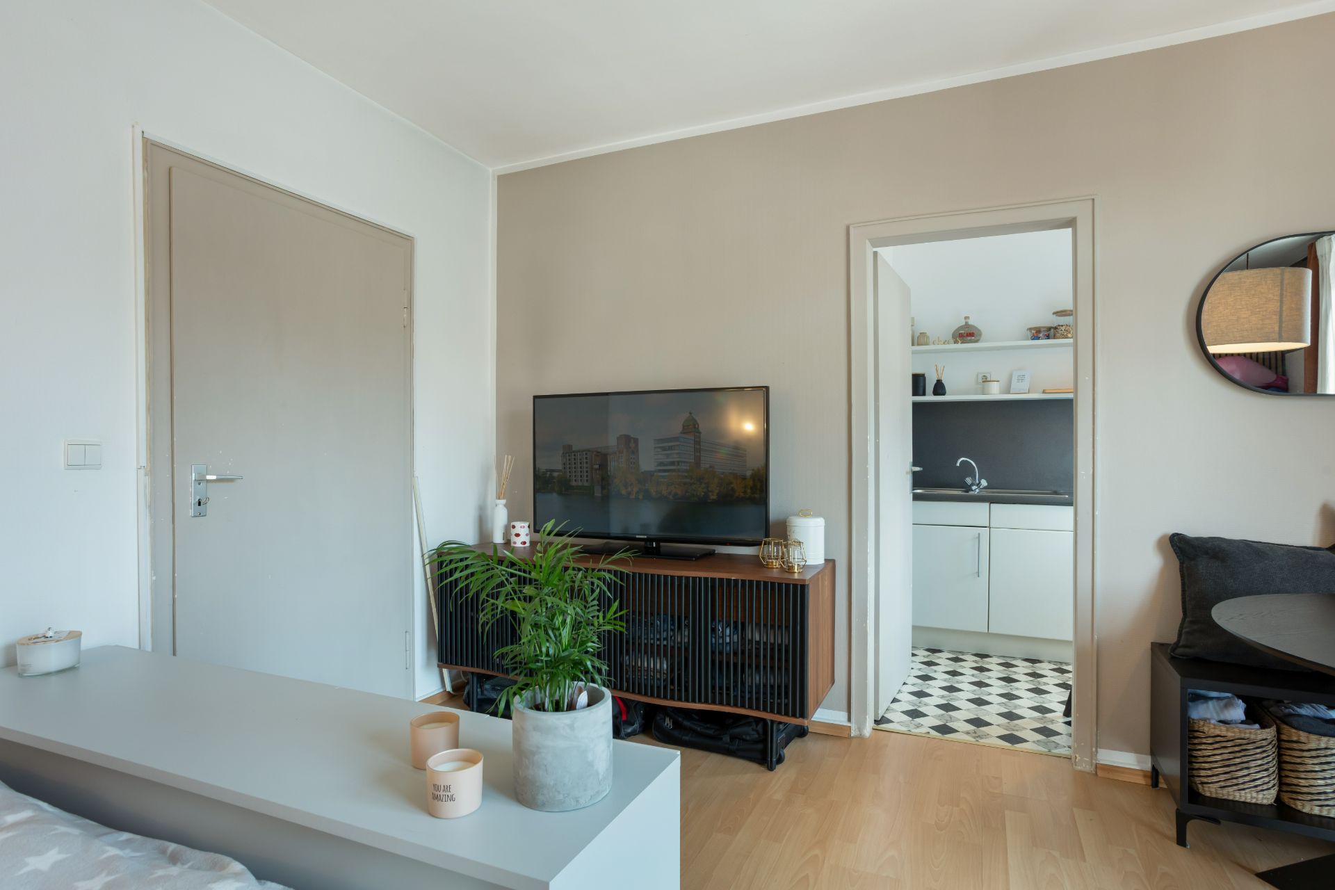 Wohnzimmer mit Fernseher, Pflanze, Tür zur weißen Küche mit schwarz-weißem Fliesenboden.