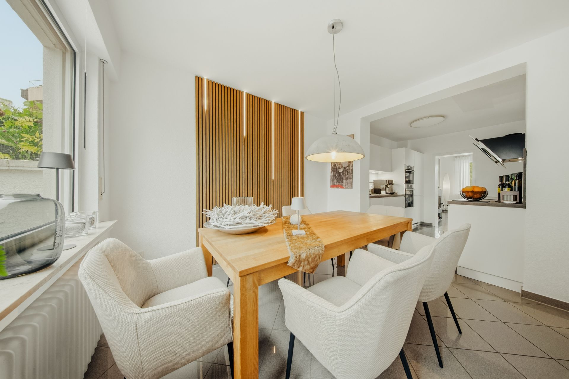 Helles Esszimmer mit Holztisch, vier weißen Stühlen und Hängelampe, angrenzend an eine offene Küche.