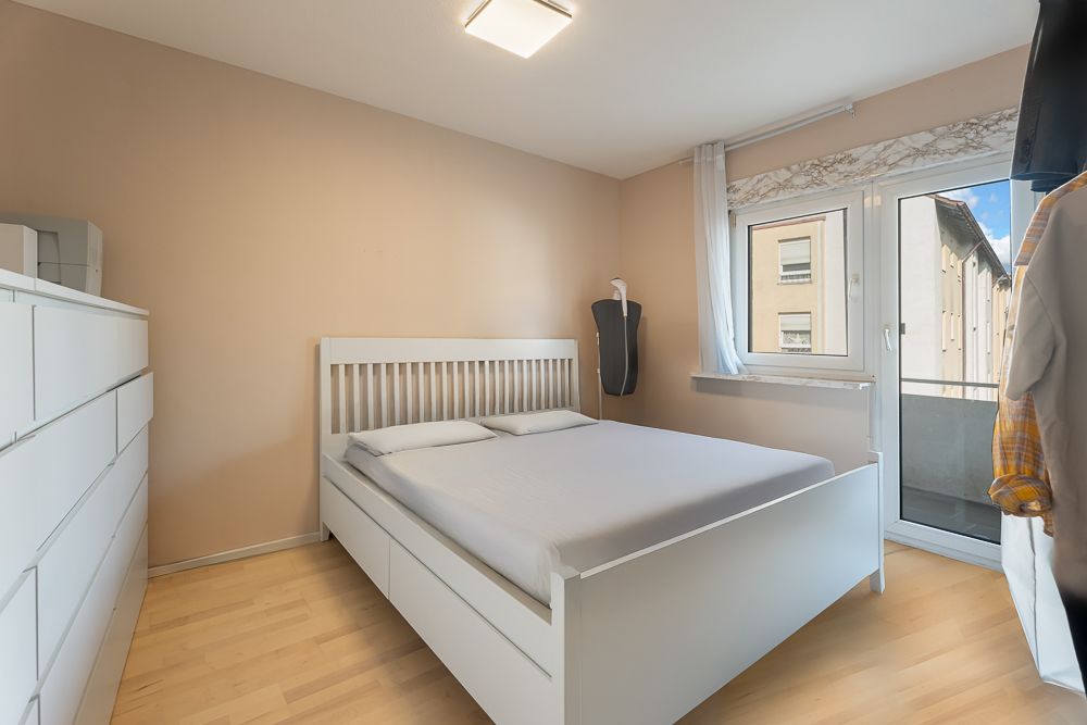 Helles Schlafzimmer mit weißem Bett, Kommode, Fenster und Zugang zu einem Balkon.