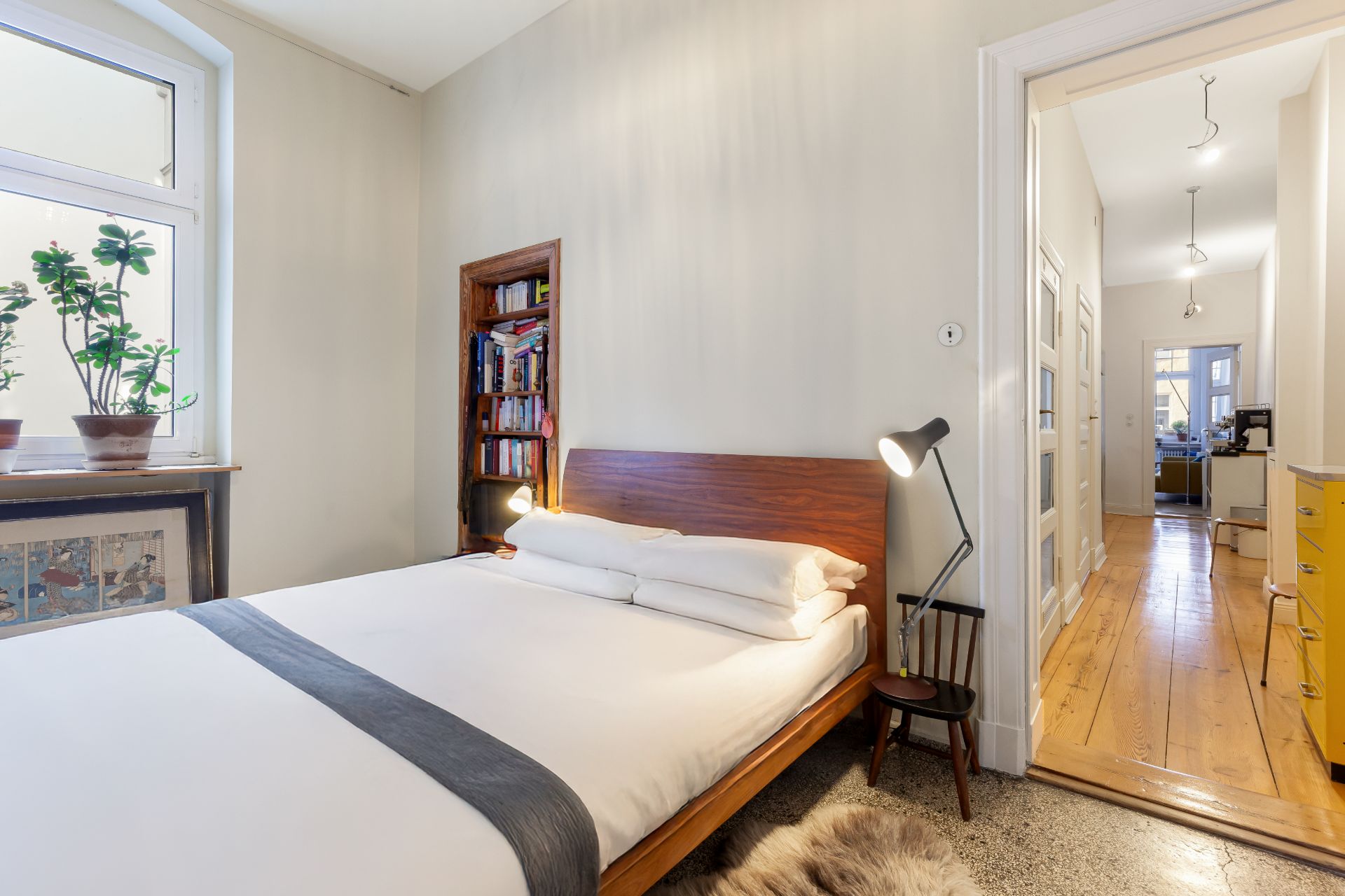 Helles Schlafzimmer mit Doppelbett, Pflanzen am Fenster, Bücherregal und Blick in Flur mit Holzboden.