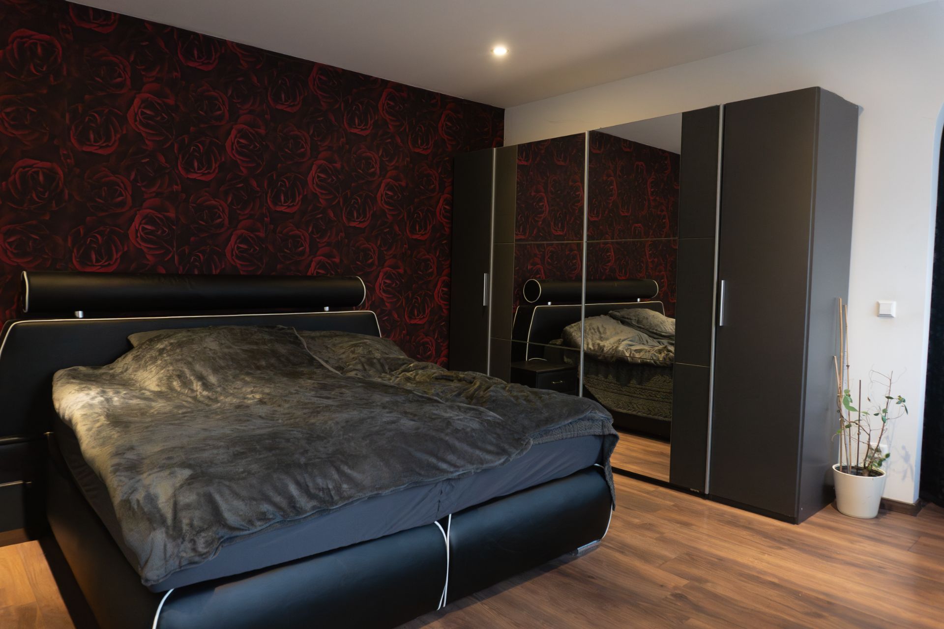 Schlafzimmer mit großem Bett, dunklem Kleiderschrank mit Spiegel und Pflanze in der Ecke.
