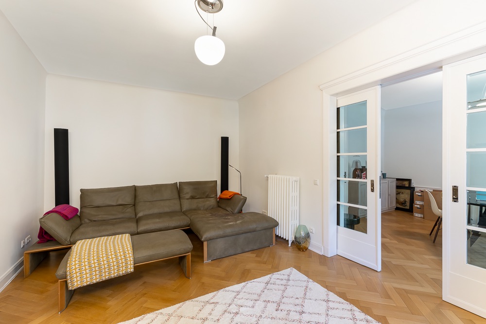 Wohnzimmer mit grauem Ecksofa, Stehlampe, Teppich und einer weißen Doppelschiebetür.