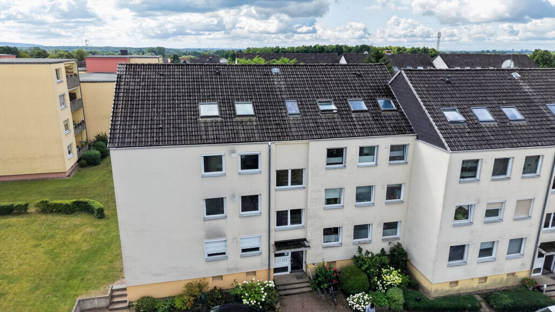 Mehrfamilienhaus mit drei Stockwerken, Satteldach und Vorgarten, umgeben von grüner Wiese.