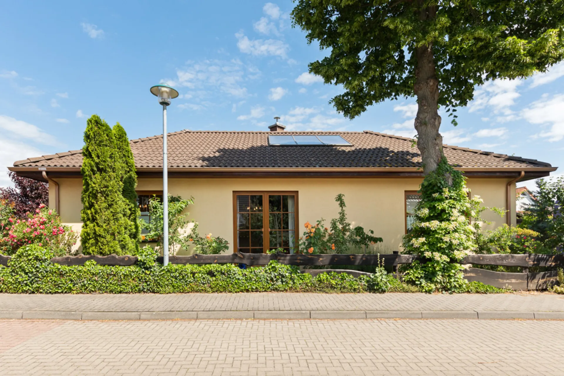 Einfamilienhaus mit Garten, Bäumen und Blumen, vor einer Straße, bei sonnigem Wetter.