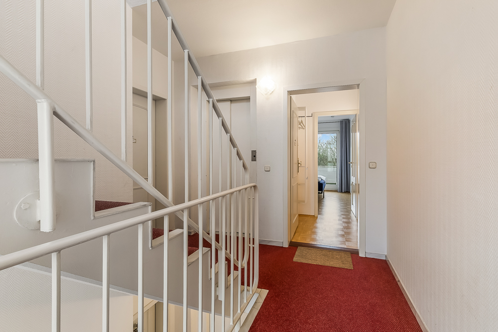 Flur mit rotem Teppich, Treppe, Aufzug und Blick in ein Zimmer mit Fenster.