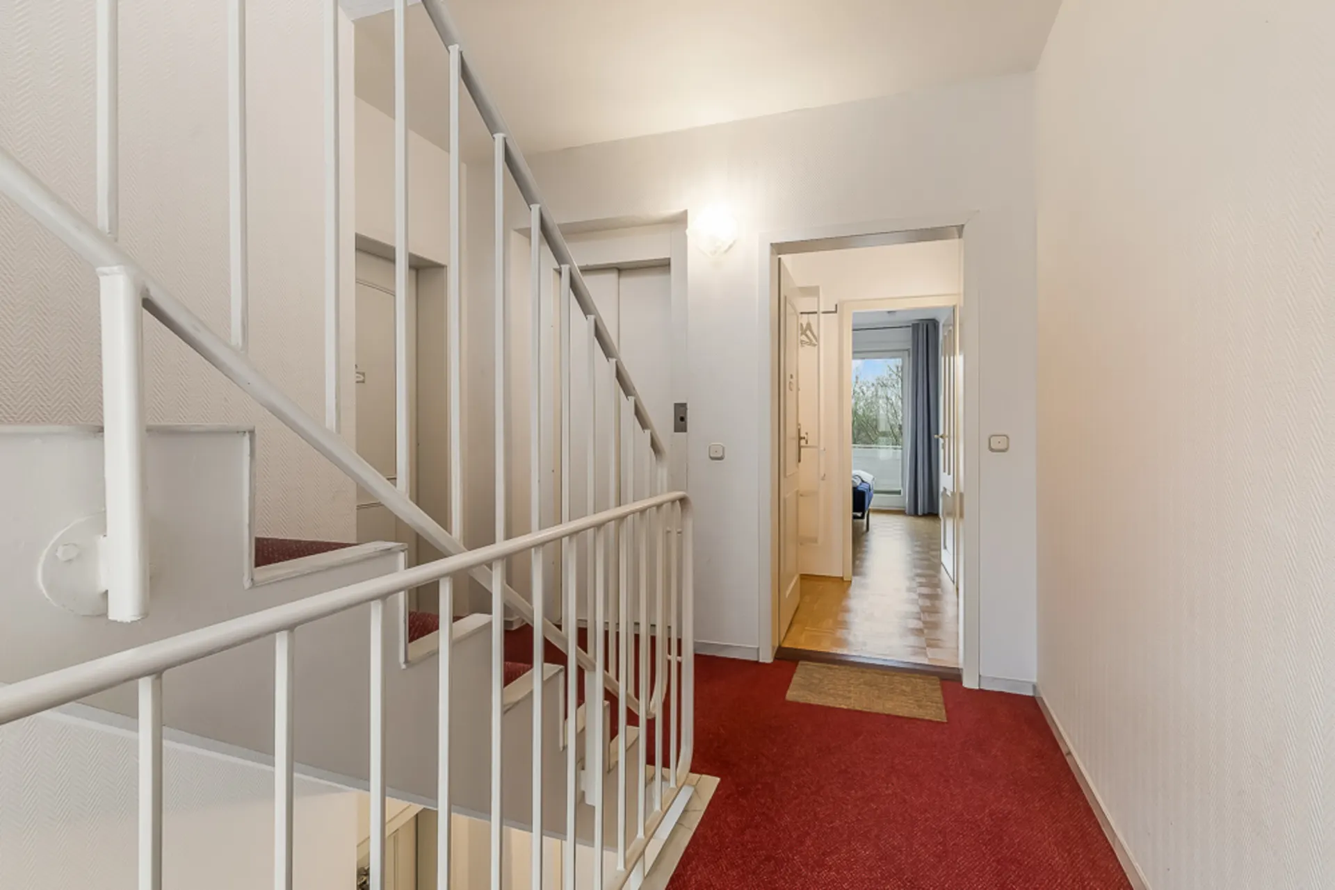 Flur mit rotem Teppich, Treppe, Aufzug und Blick in ein Zimmer mit Fenster.