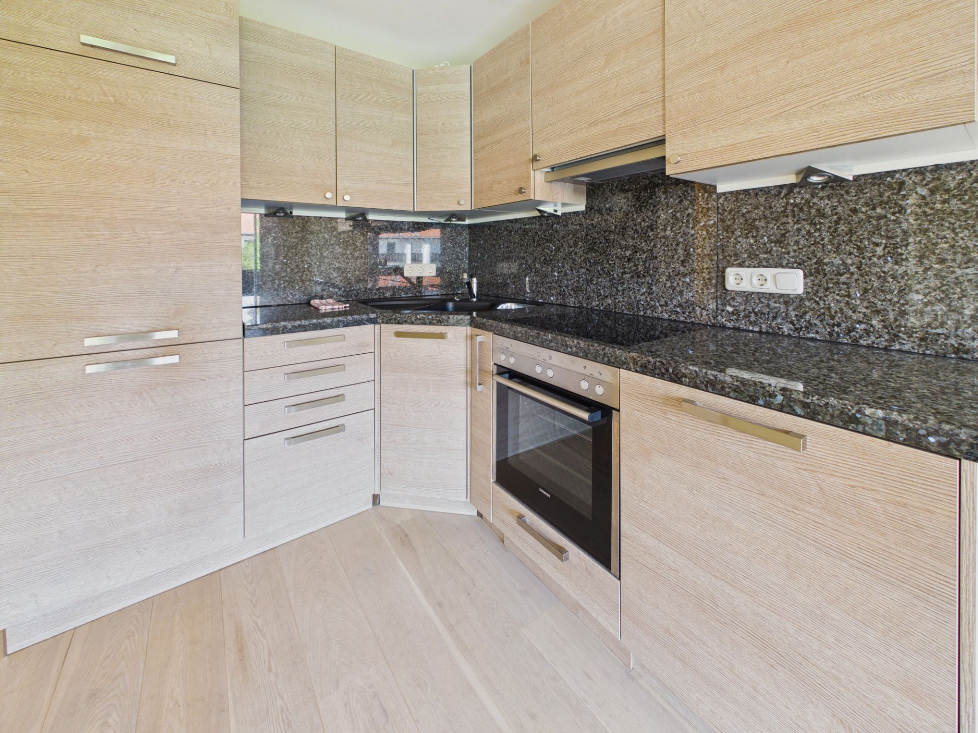 Moderne Küche mit hellen Holzmöbeln, schwarzer Granit-Arbeitsplatte und integriertem Backofen.