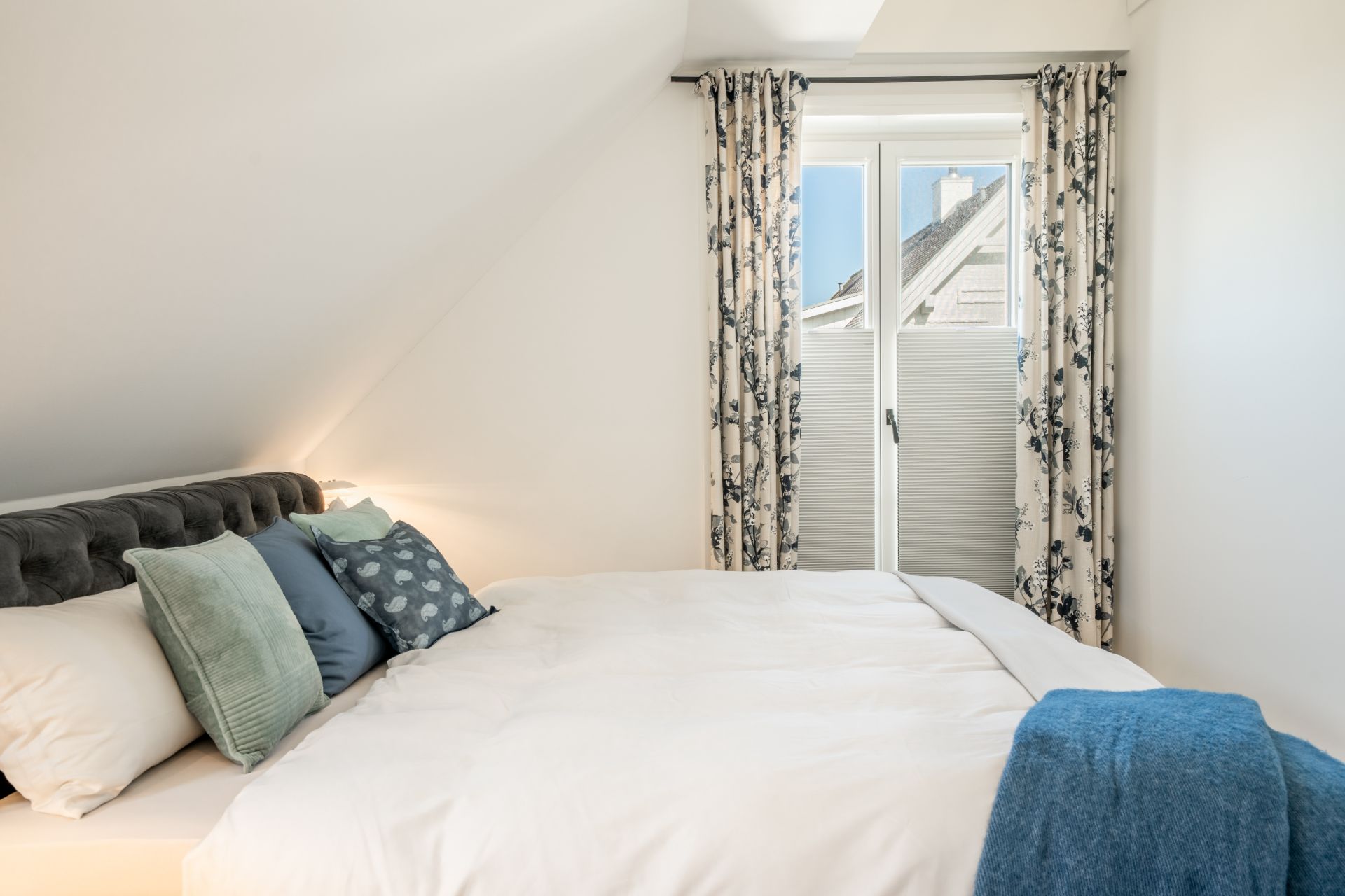 Helles Schlafzimmer mit Doppelbett, bunten Kissen und Fenster mit gemusterten Vorhängen.
