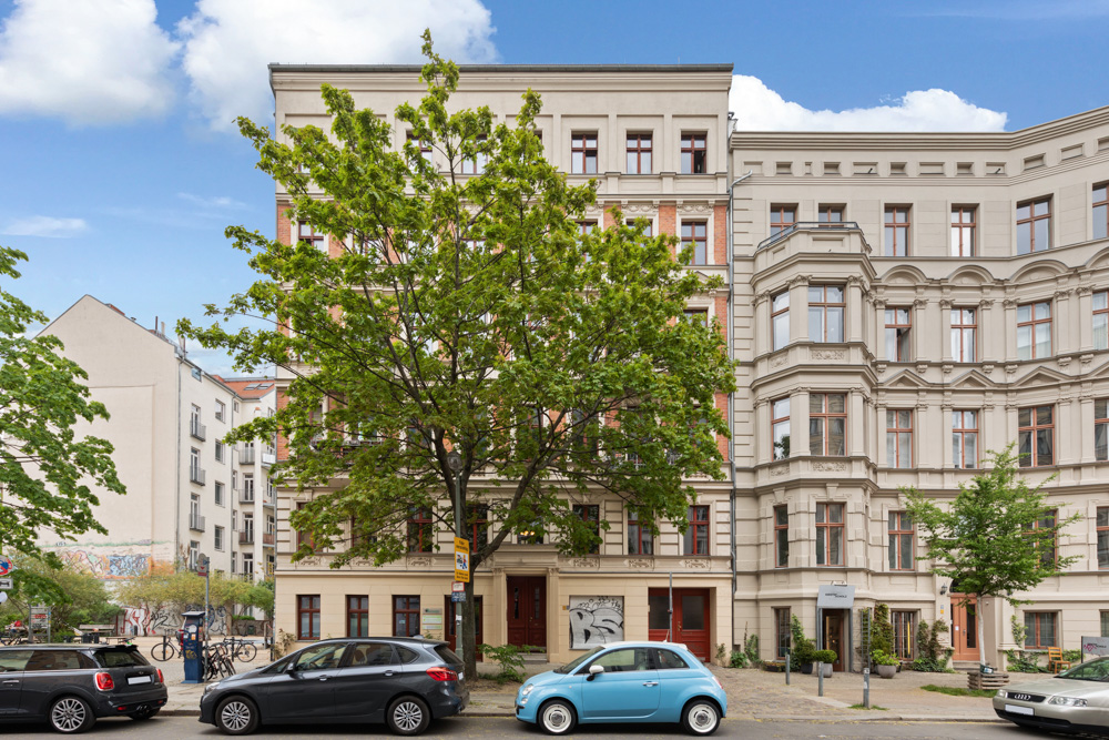 Mehrstöckiges Wohnhaus mit großen Fenstern, Bäumen und parkenden Autos davor.