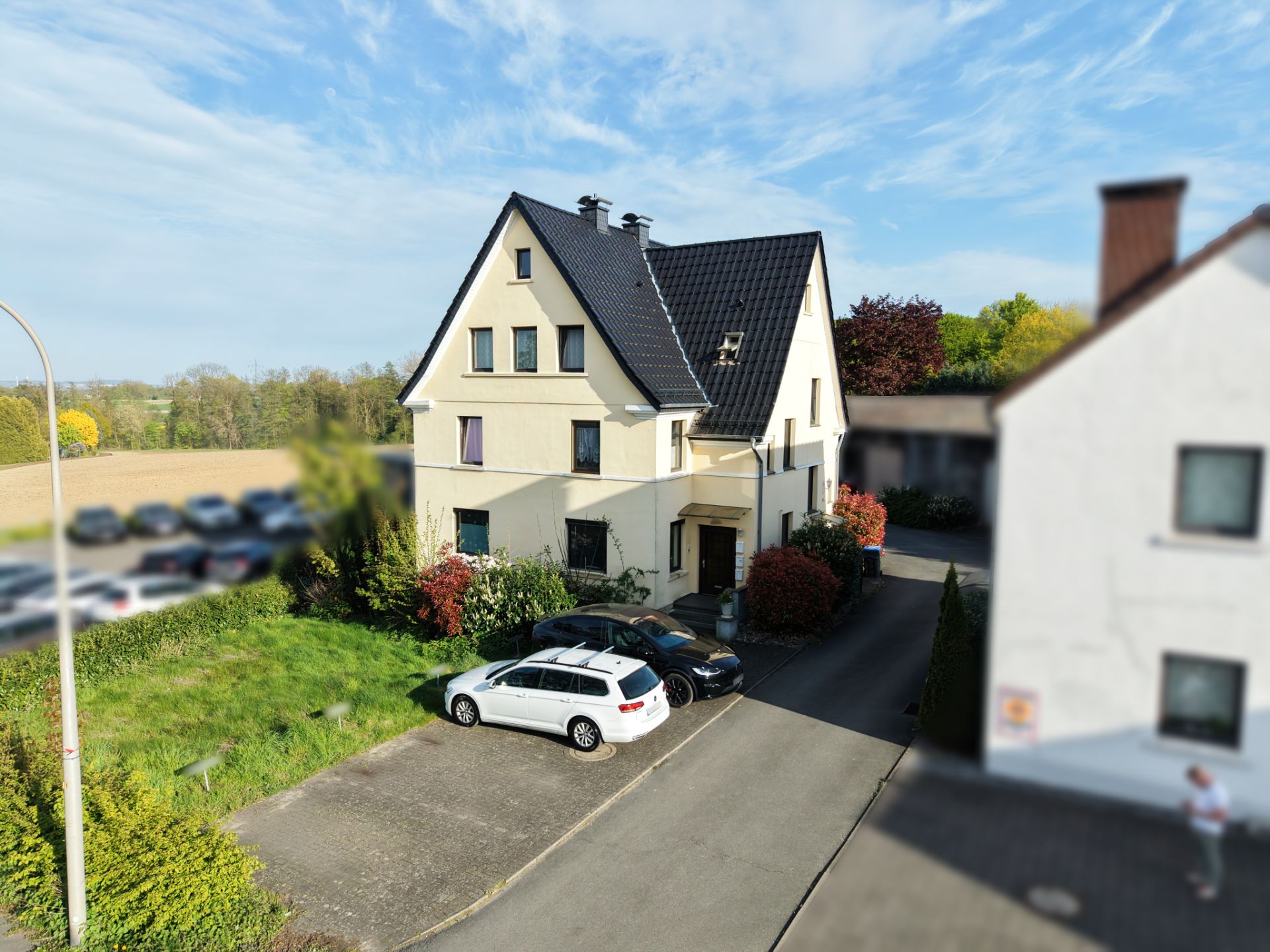 Zweistöckiges Einfamilienhaus mit schwarzem Dach, davor zwei parkende Autos und einige Büsche.