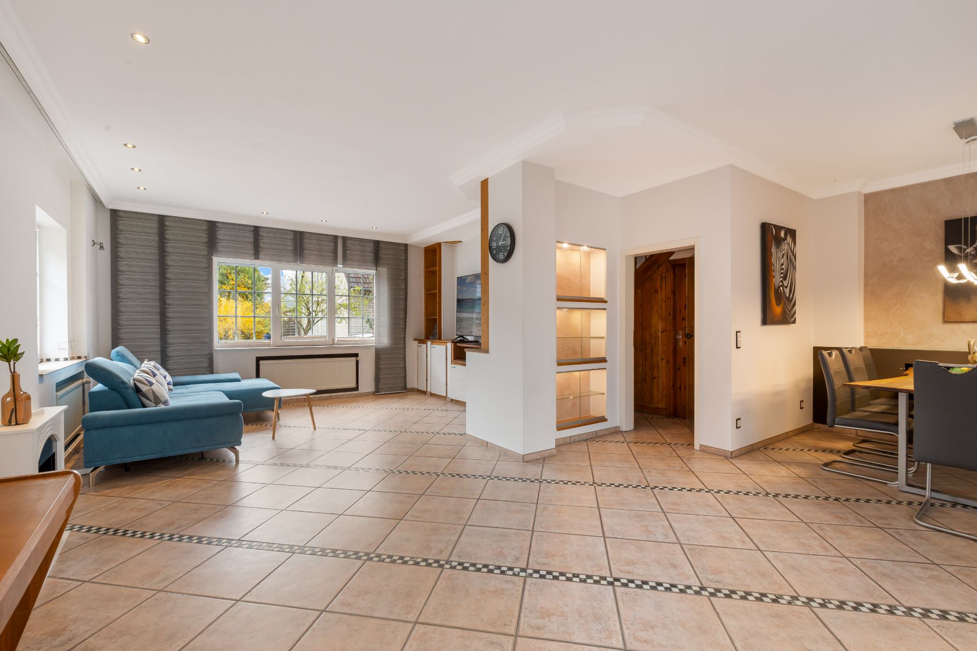 Großes Wohnzimmer mit Sofa, Esstisch, Fliesenboden und großem Fenster mit Blick ins Freie.