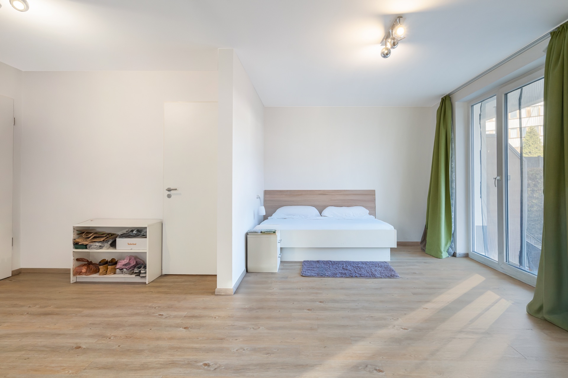 Helles Schlafzimmer mit Doppelbett, Nachttisch, Teppich und großem Fenster mit grünen Vorhängen.