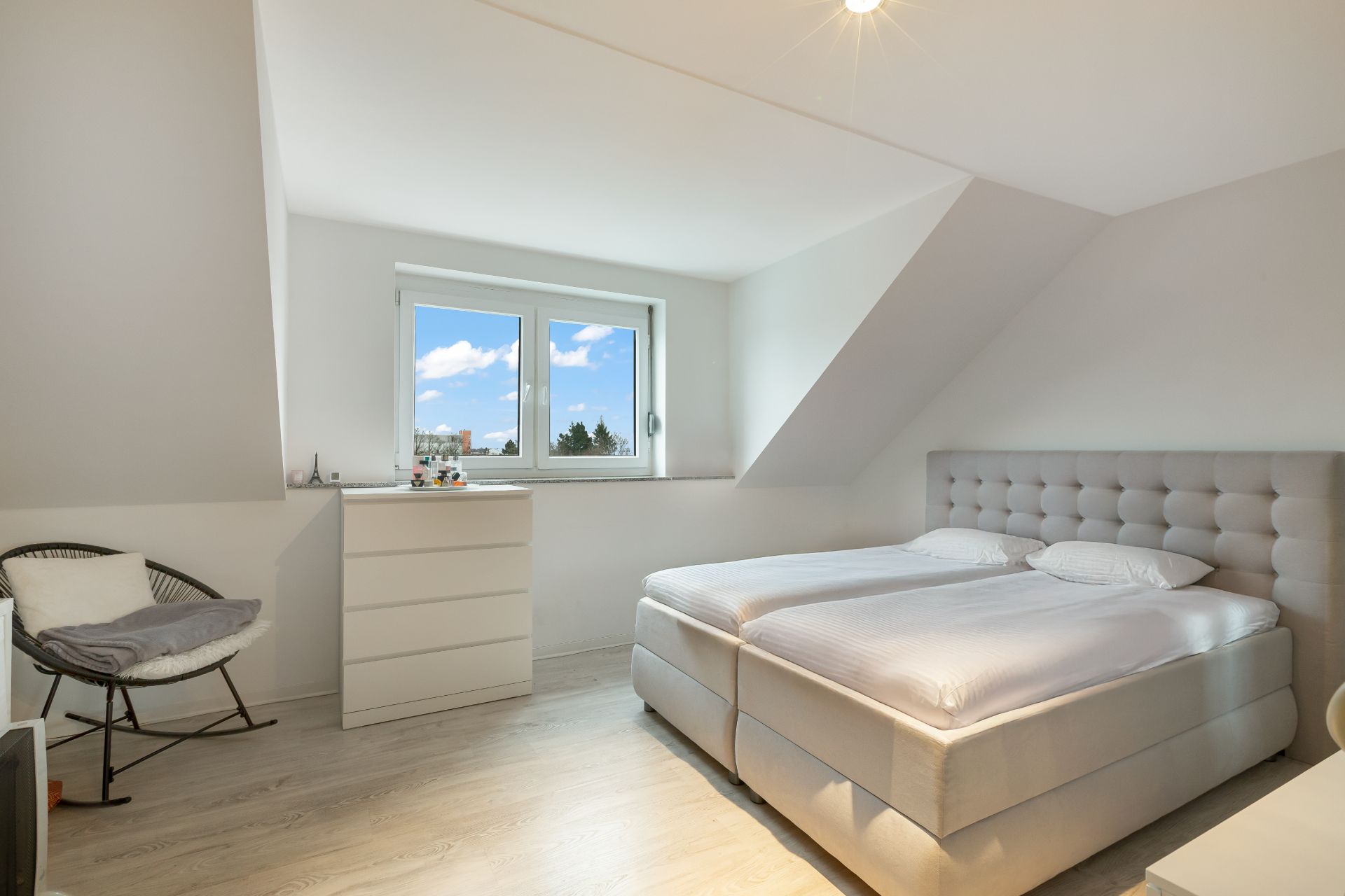 Helles Schlafzimmer mit Doppelbett, Kommode und Stuhl unter einem Dachfenster.