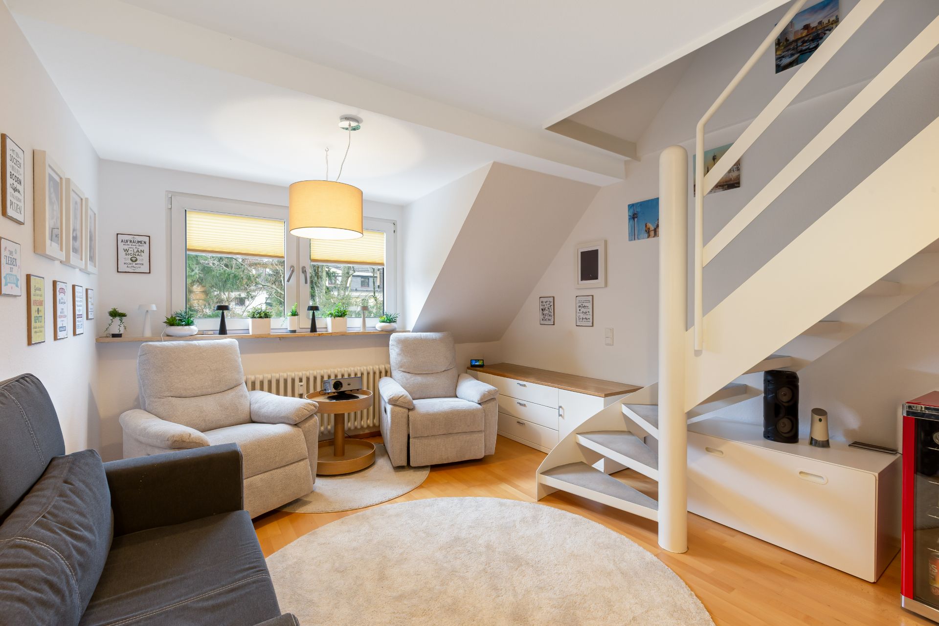 Helles Wohnzimmer mit Sofa, zwei Sesseln, runder Teppich und Treppe nach oben.