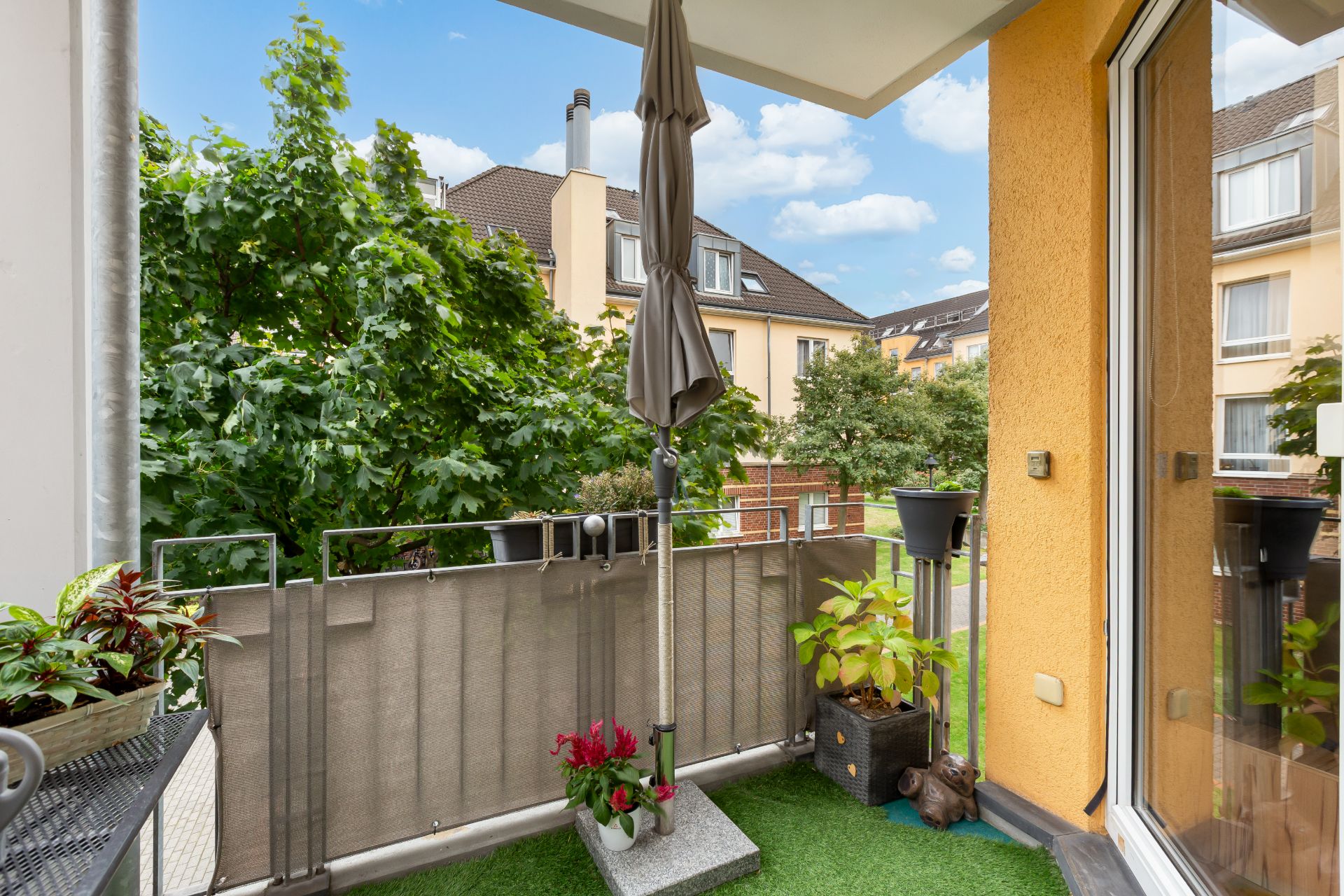Balkon mit Pflanzen, Sonnenschirm und Blick auf Bäume und benachbarte Wohnhäuser.