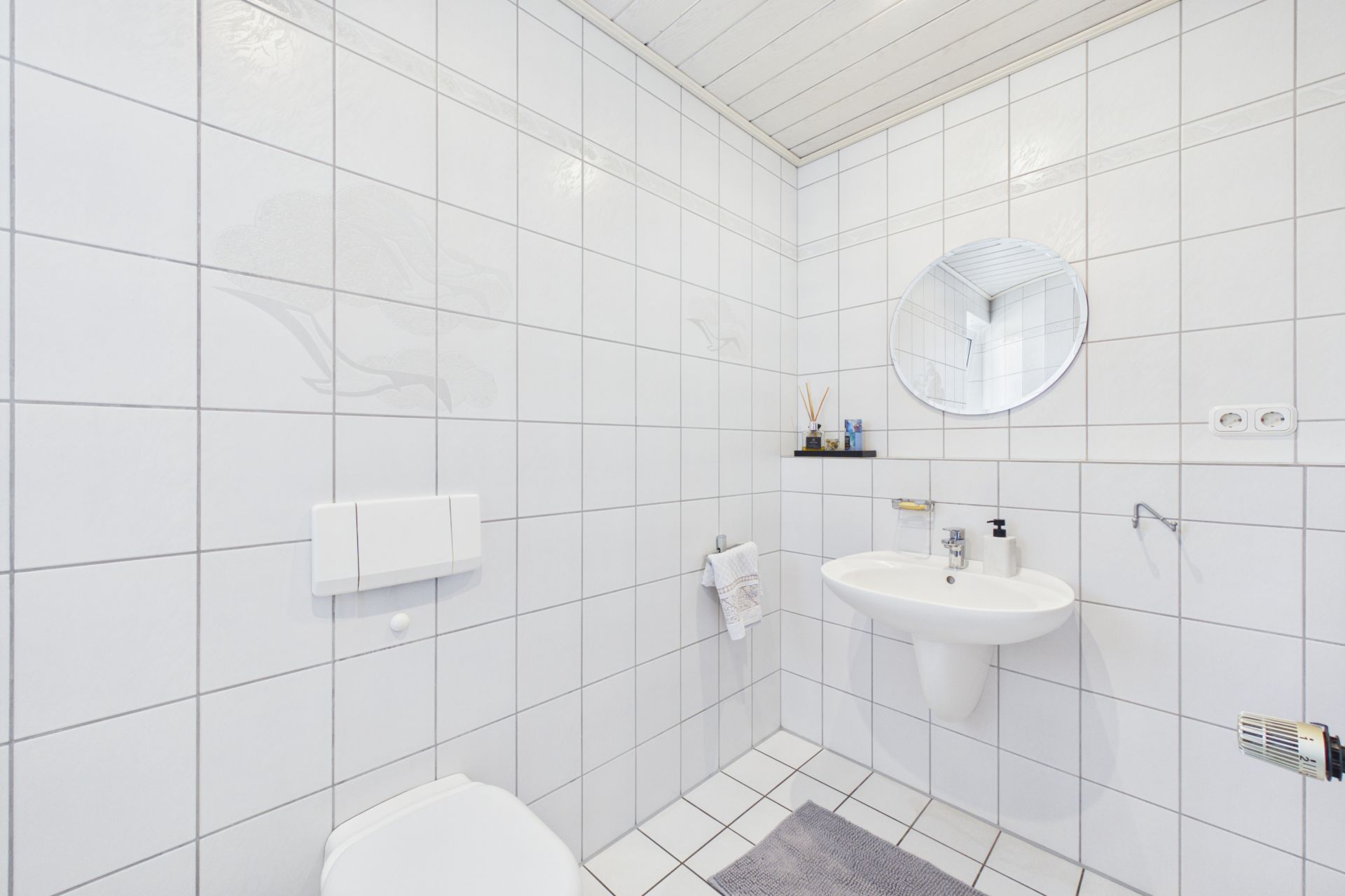 Helles Badezimmer mit weiß gefliesten Wänden, Waschbecken, rundem Spiegel und Toilette.
