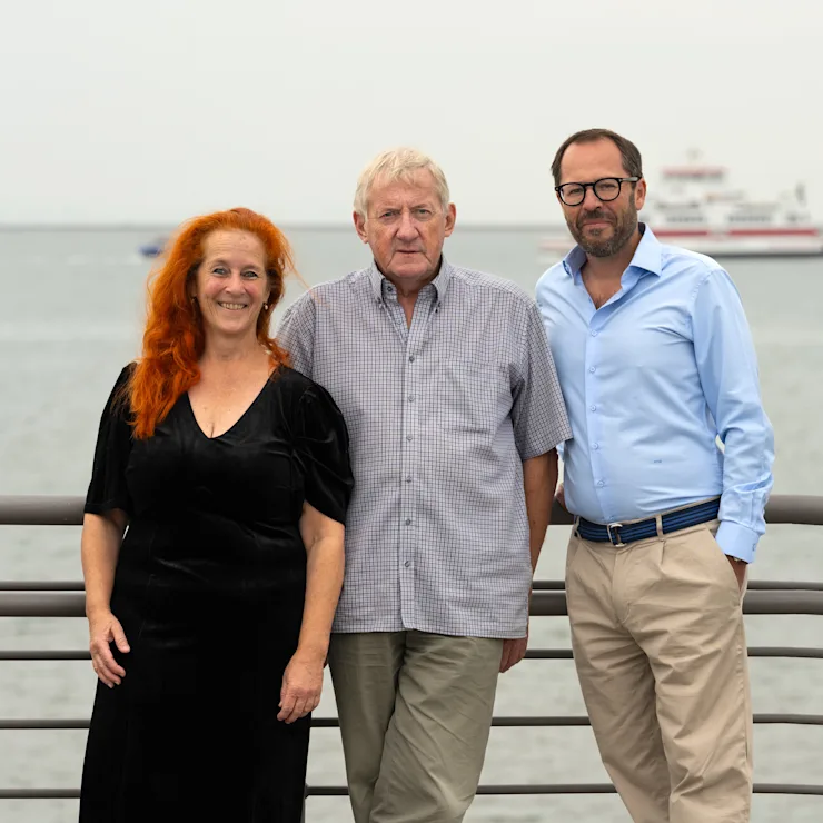 Drei Erwachsene stehen an einem Geländer mit Wasser und Schiff im Hintergrund.