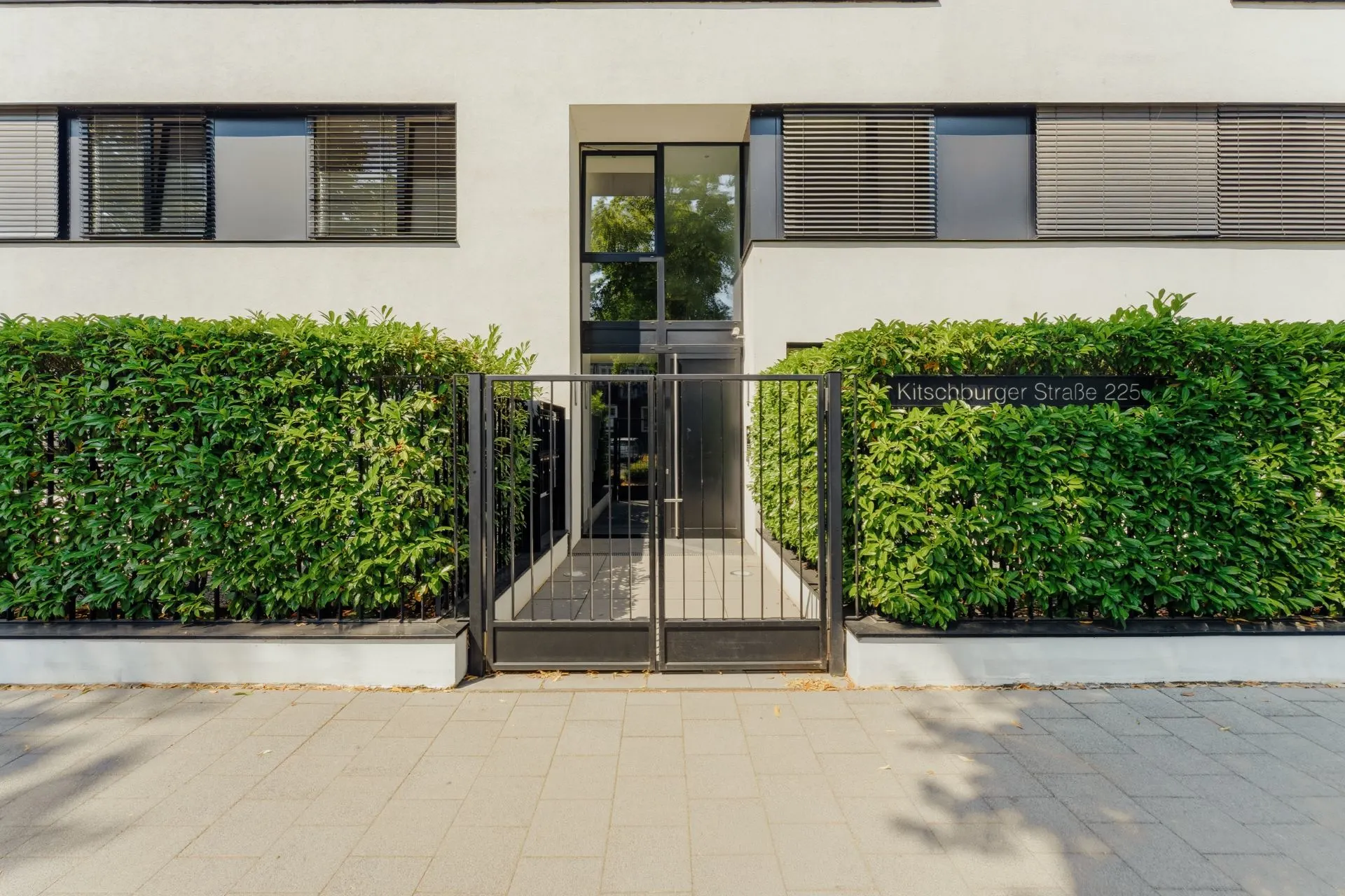 Eingang eines modernen Wohnhauses mit Metalltor und Hecke an der Straße.