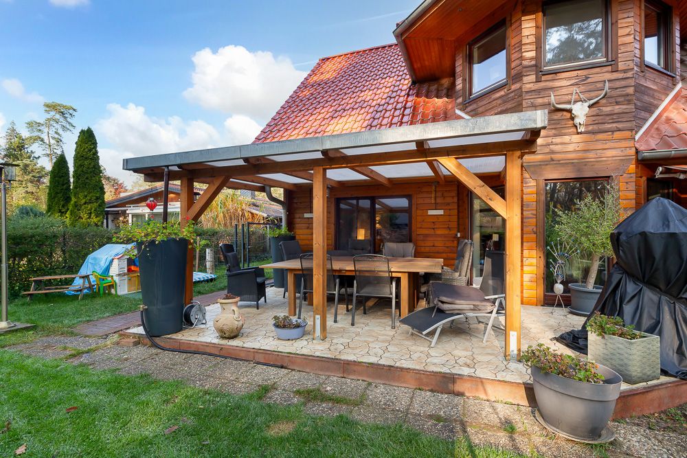 Überdachte Terrasse mit Holztisch und Stühlen an einem Holzhaus, umgeben von Pflanzen im Garten.