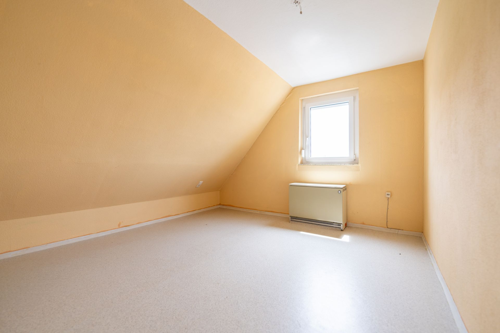 Leeres Zimmer mit schräger Decke, gelben Wänden und einem Fenster mit Heizkörper darunter.