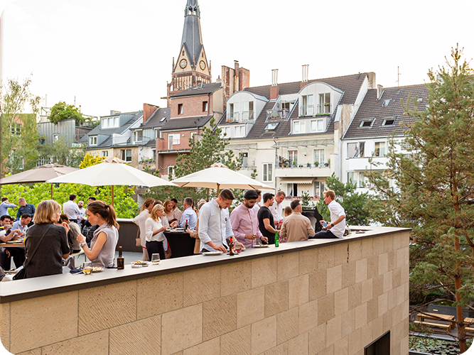 Menschen feiern auf einer Dachterrasse mit Sonnenschirmen, im Hintergrund Wohnhäuser und Bäume.