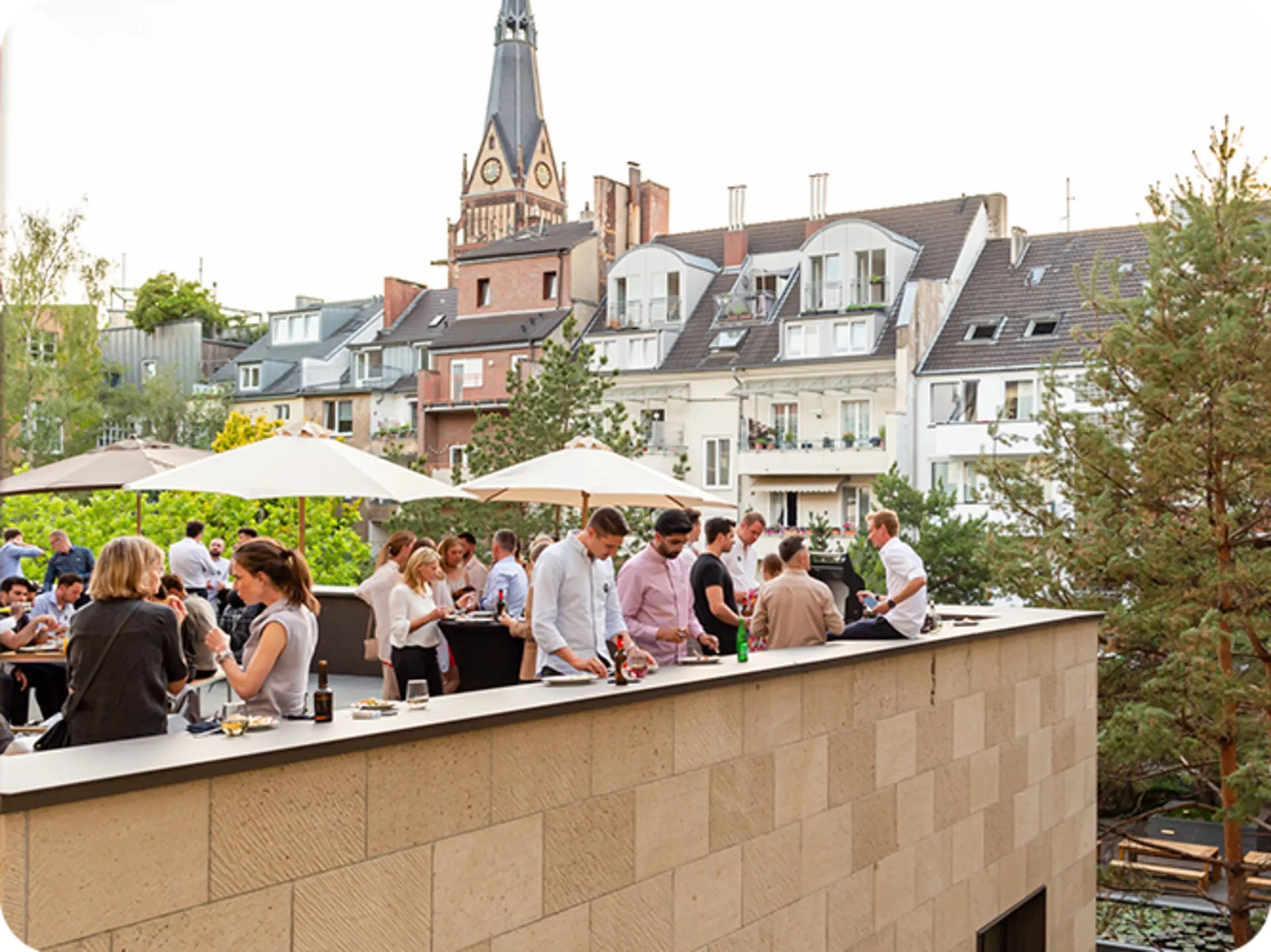 Menschen feiern auf einer Dachterrasse mit Sonnenschirmen, im Hintergrund Wohnhäuser und Bäume.