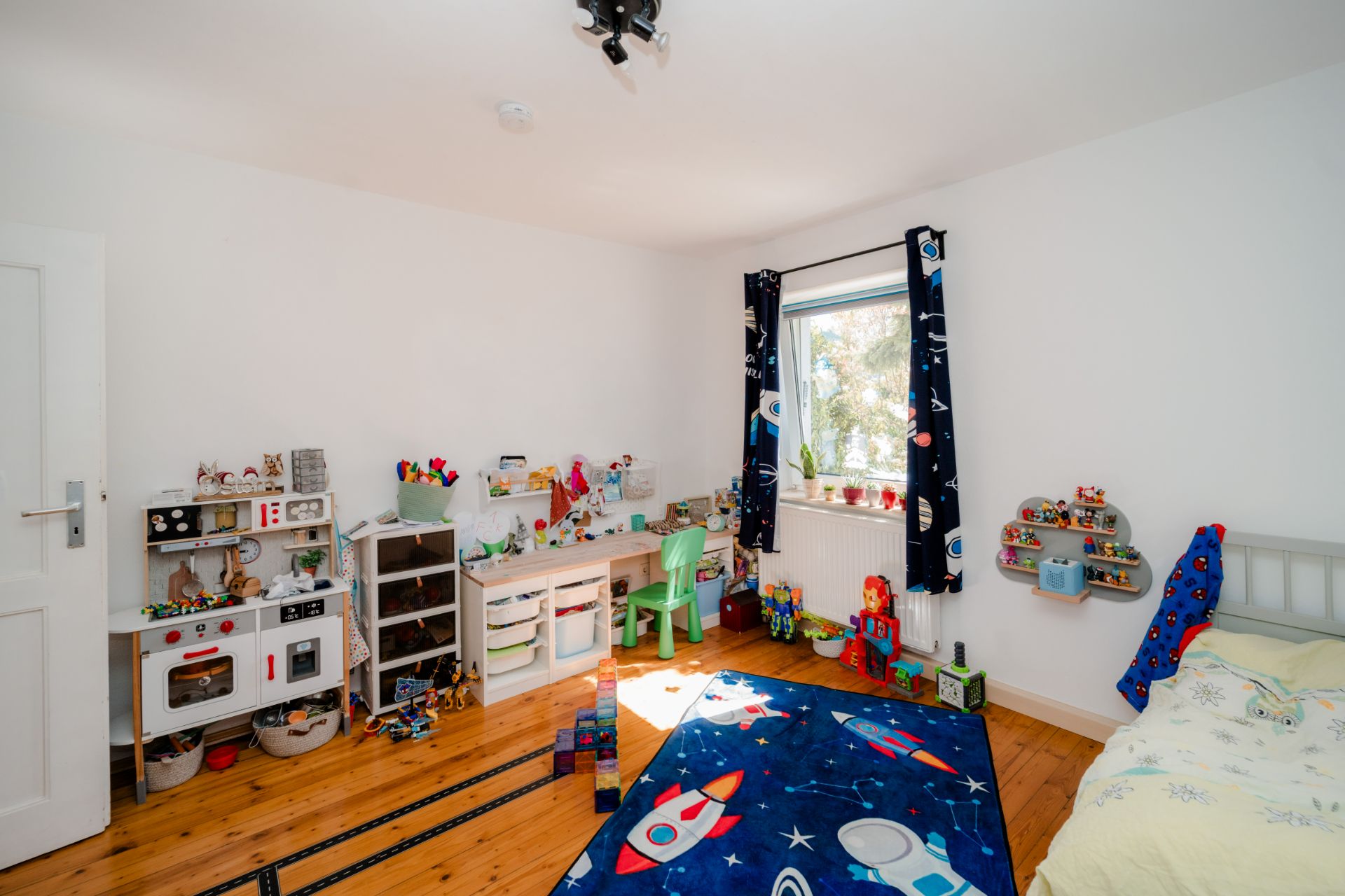 Helles Kinderzimmer mit Spielzeug, Schreibtisch, Regal, buntem Teppich und Einzelbett.