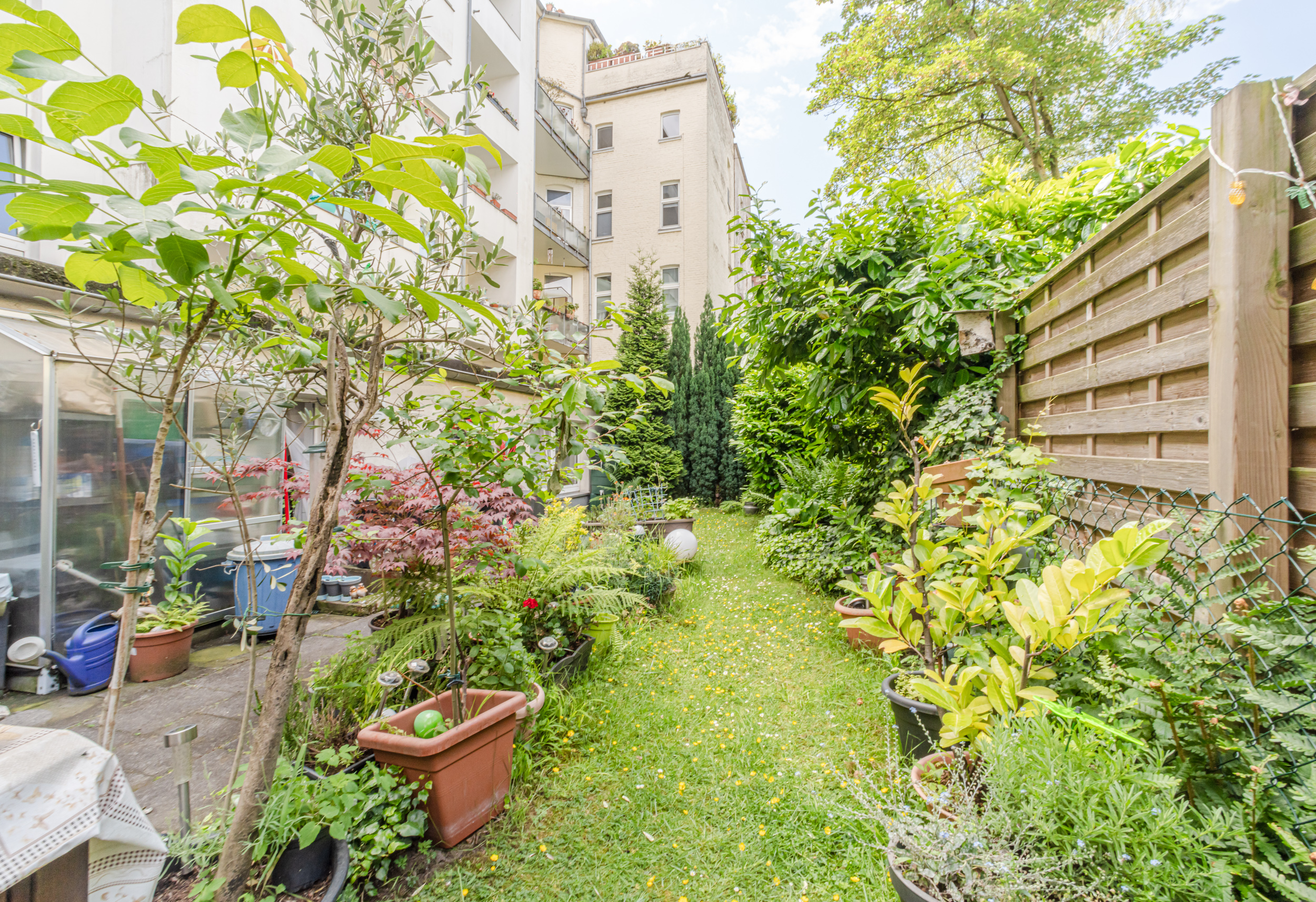Schmaler Garten mit vielen Topfpflanzen, Bäumen und Sträuchern neben einem Wohnhaus.