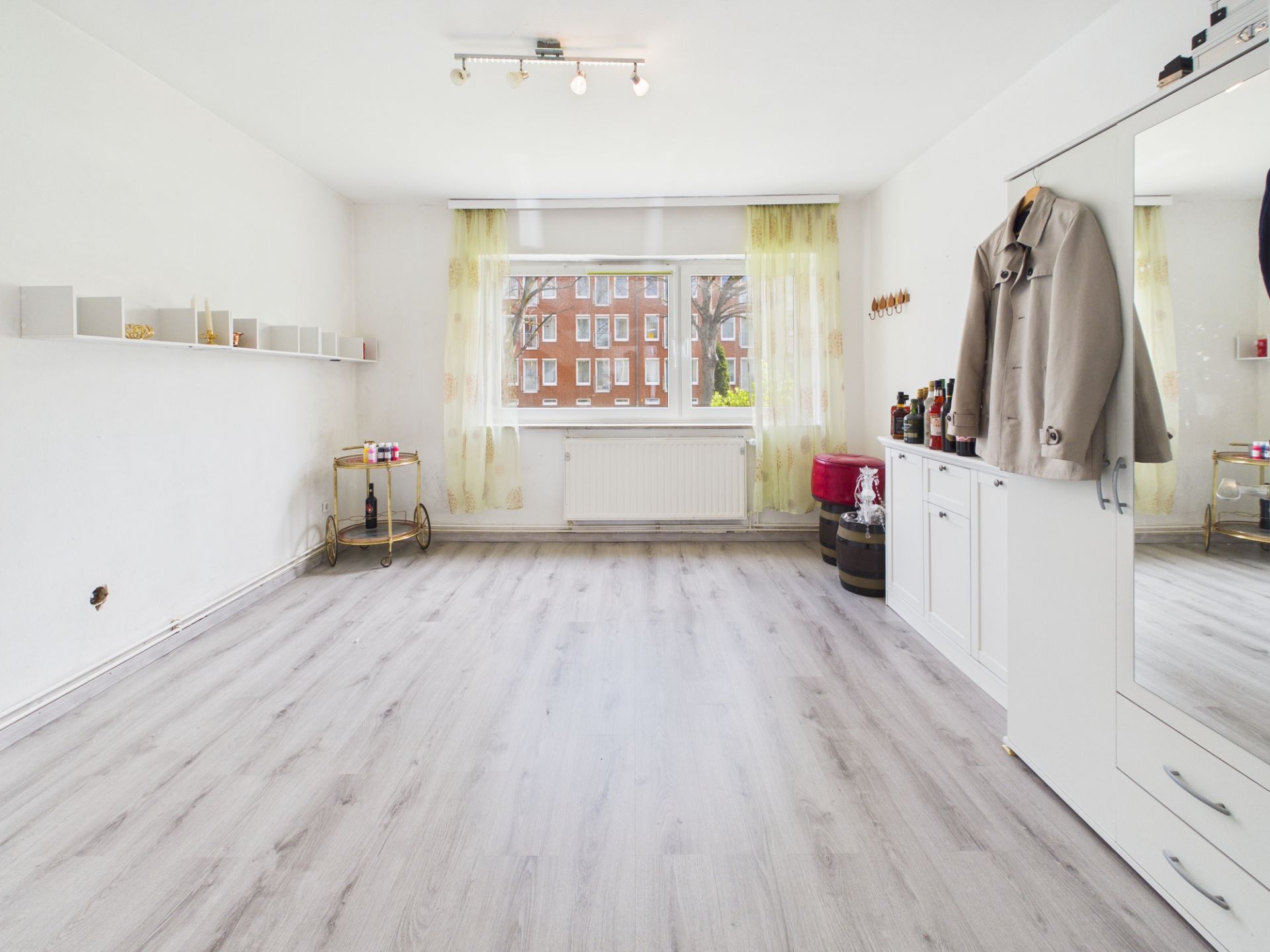 Helles Zimmer mit großem Fenster, Laminatboden, weißem Schrank und Wandregalen.