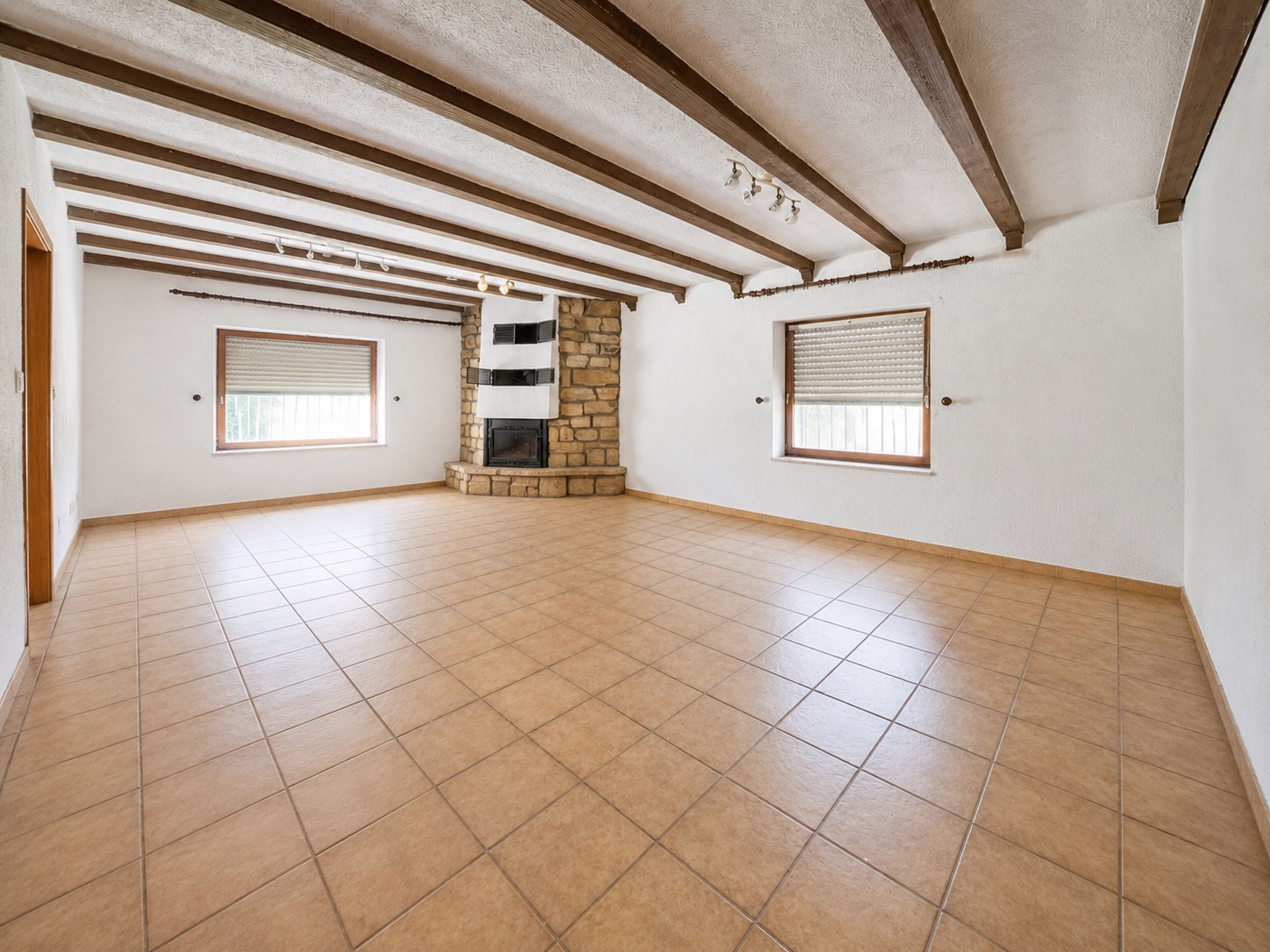 Großes, leeres Wohnzimmer mit Fliesenboden, Holzbalken und Kamin aus Stein.