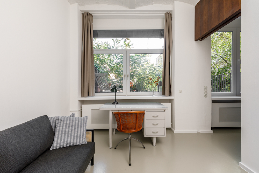 Helles Zimmer mit großem Fenster, Schreibtisch und Stuhl, daneben ein Sofa mit Kissen.