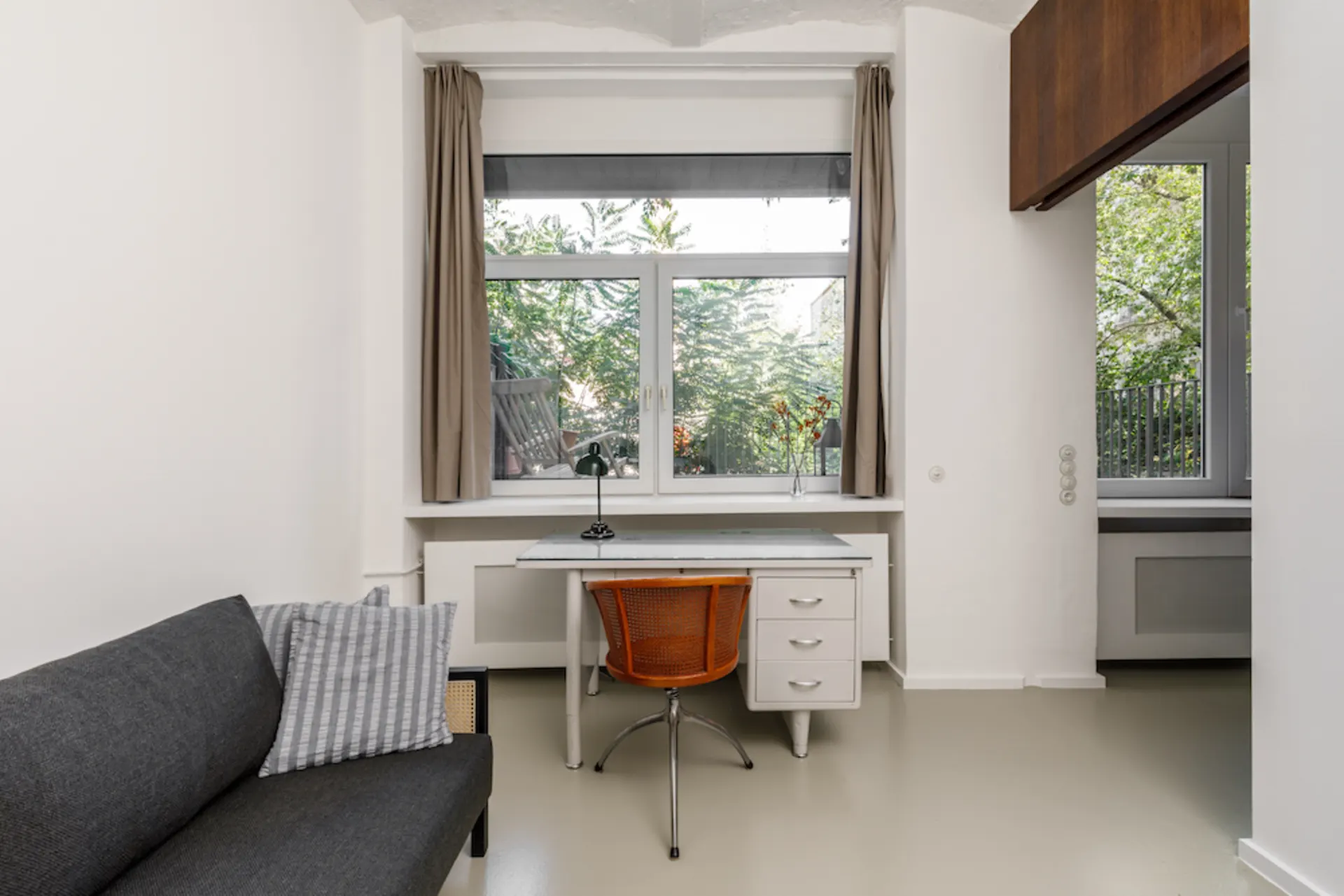 Helles Zimmer mit großem Fenster, Schreibtisch und Stuhl, daneben ein Sofa mit Kissen.