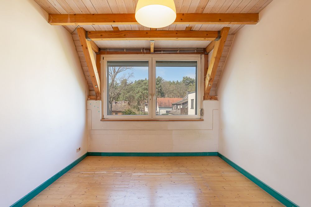 Leeres Zimmer mit Holzdecke, großem Fenster und Blick auf Bäume und Dächer.