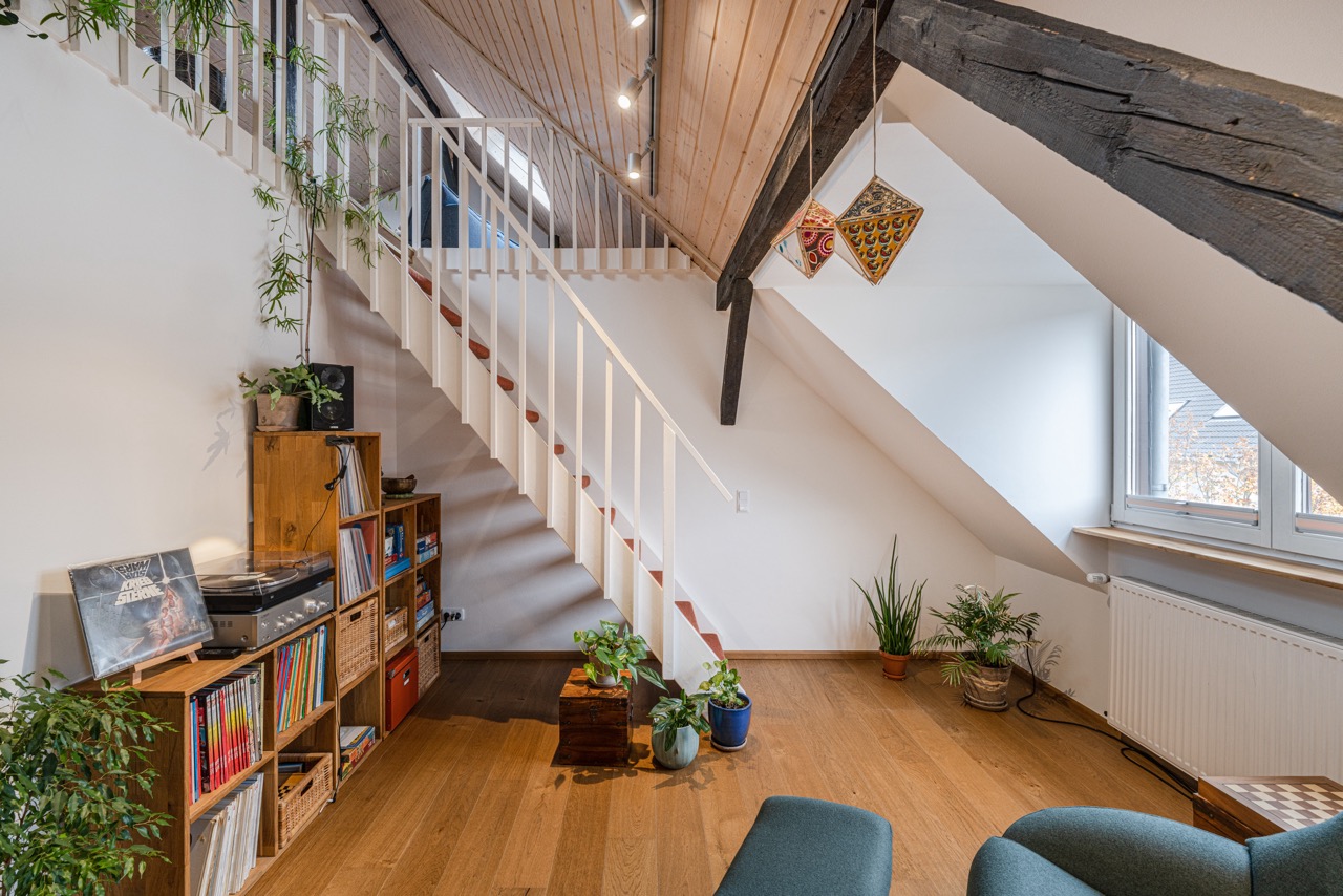 Wohnzimmer mit Holztreppe, Bücherregal, Pflanzen und schräger Decke.