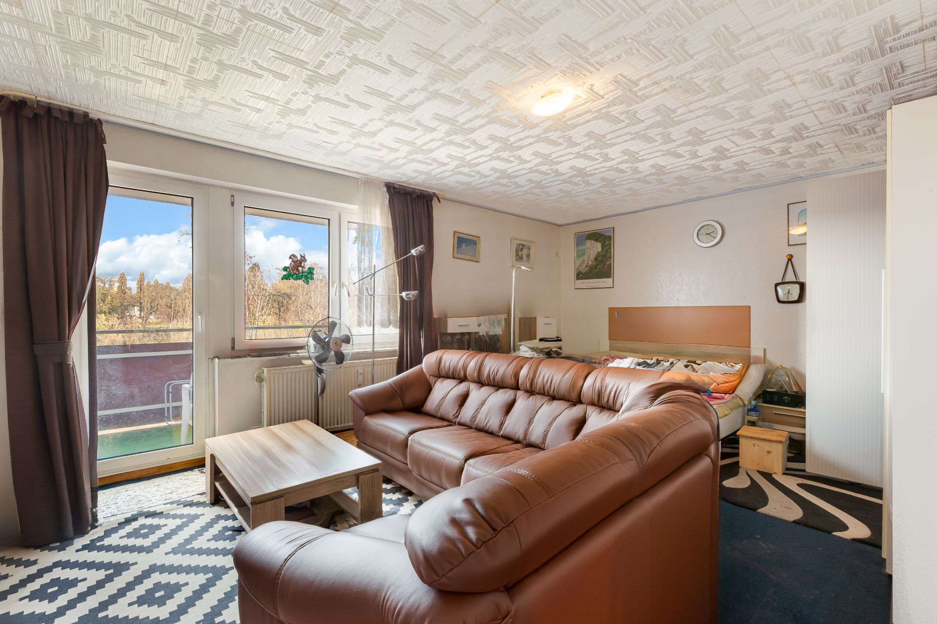 Wohnzimmer mit braunem Sofa, Couchtisch, Bett und Balkonzugang mit großen Fenstern.