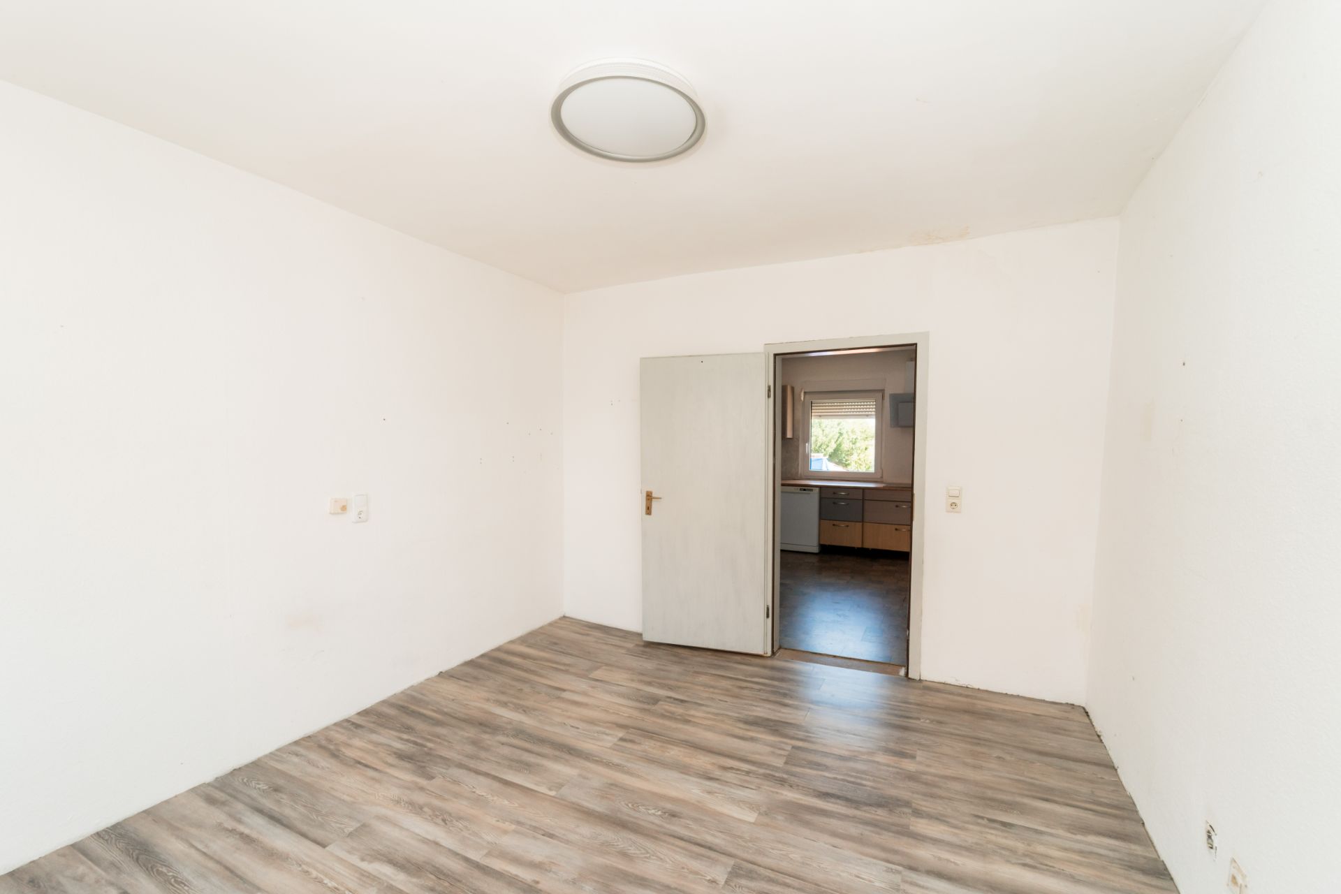 Leeres Zimmer mit Holzfußboden und weiß gestrichenen Wänden, Tür führt in einen anderen Raum mit Fenster.